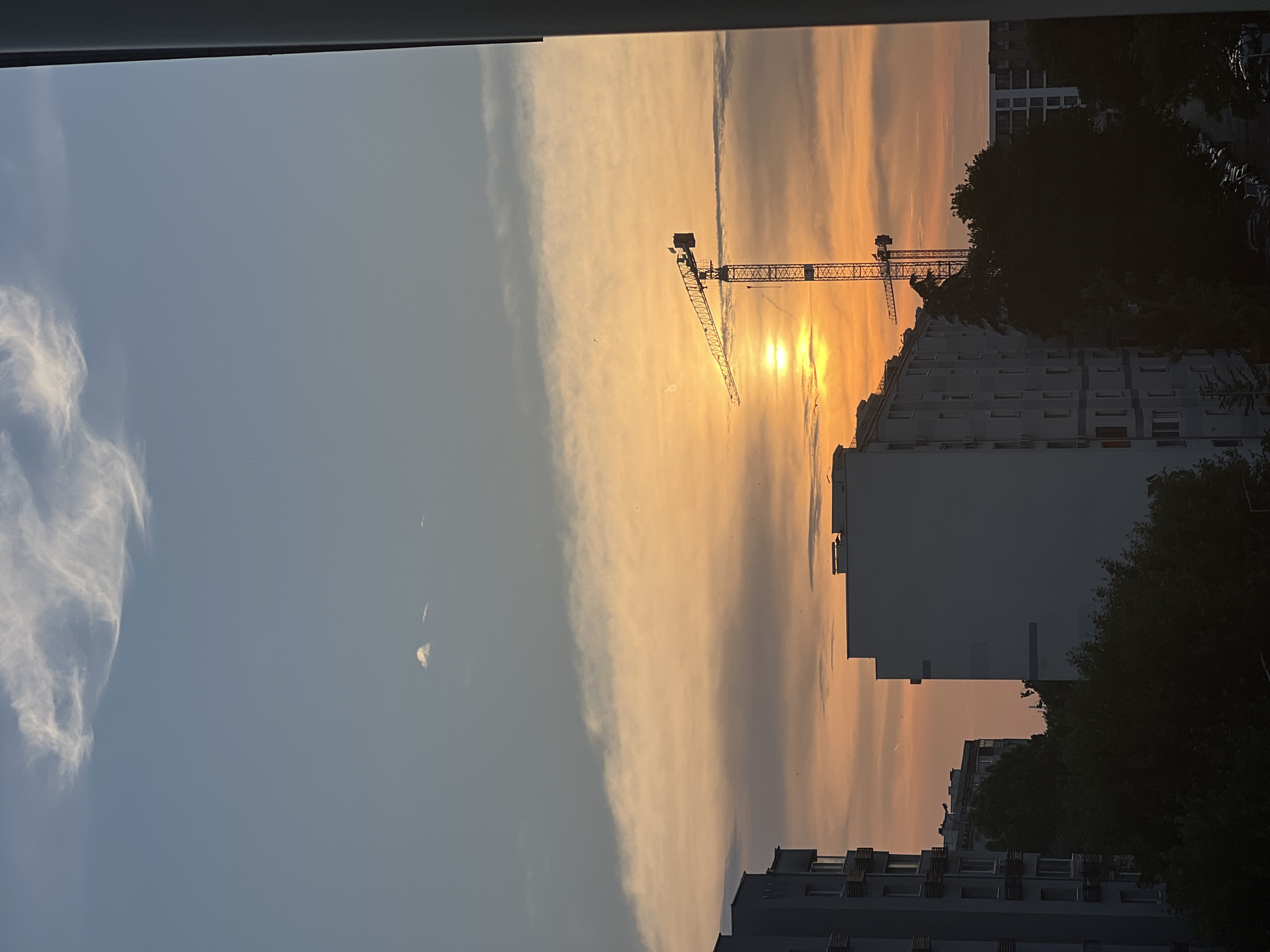 The image depicts an urban sunset scene, specified as being in the city of Burgas, Bulgaria. The upper portion of the frame features a pale blue sky transitioning into warm orange, yellow, and pink hues closer to the horizon. Wispy white clouds are present in the upper left, with a distinct long, horizontal cloud band stretching across the mid-section, colored by the setting sun. The sun itself is a bright, indistinct disc positioned in the lower-right quadrant of the sky, partially obscured by atmospheric haze.
Several multi-story residential buildings dominate the middle and lower sections of the image. On the left, a light-colored apartment building with numerous windows and balconies is partially visible. Towards the center-right, another large, pale building stands, silhouetted against the bright sky. A prominent yellow construction tower crane is centrally located, its metallic structure creating a dark silhouette directly in front of the sun. Dark green trees fill the mid-ground, casting deep shadows that obscure their details. Below the tree line, a street with multiple parked vehicles is faintly discernible in deep shadow. The extreme right edge of the image shows a dark vertical structure, indicating an interior architectural element like a window frame or balcony edge from the vantage point of the photographer. No individuals are visible, and no specific actions or interactions are discernible. The overall environment is one of an active urban landscape at dusk.