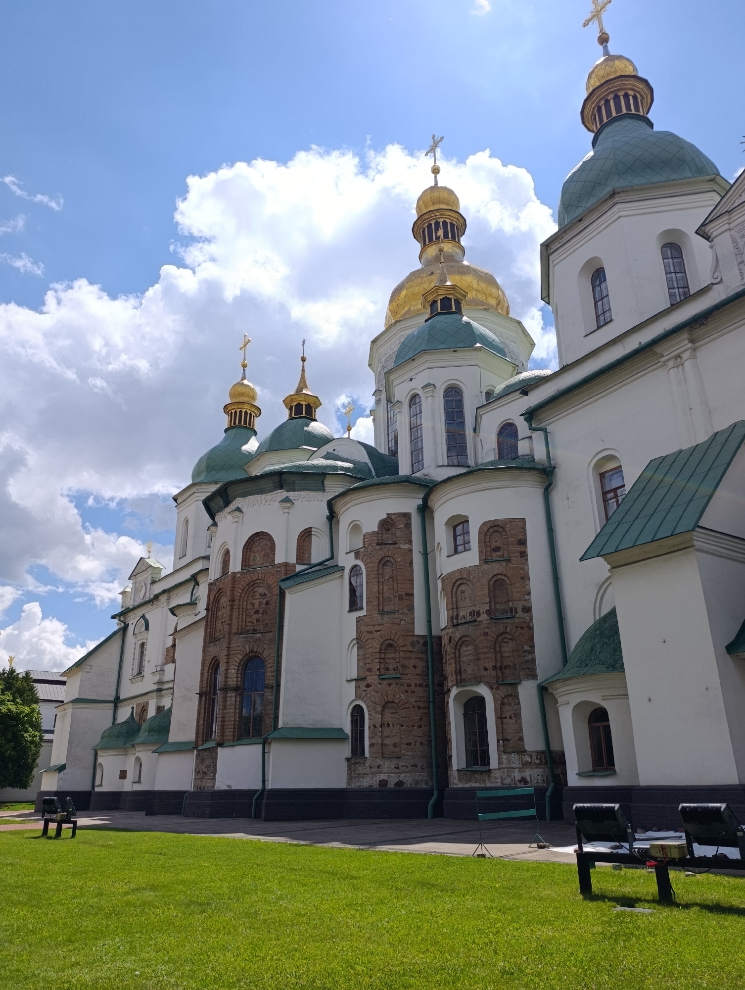 The image shows the exterior of Saint Sophia's Cathedral in Kyiv, Ukraine, on a sunny day with scattered clouds. The cathedral, a UNESCO World Heritage site, features white walls, green domed roofs, and golden spires topped with crosses. The architecture is a blend of Baroque and Byzantine styles. In the foreground, a well-maintained green lawn is visible, with a few benches and lighting equipment placed on a paved area at the edge of the grass. The perspective is from a low angle, looking up at the grand structure, emphasizing its height and the vibrant sky above.