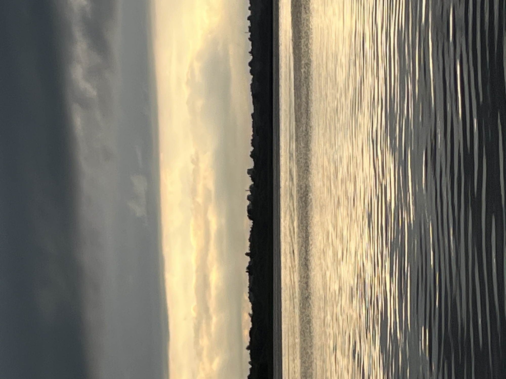The image depicts a wide body of water with rippling surface, reflecting the sky above. A dark, silhouetted treeline with a few buildings is visible on the horizon. The sky is characterized by distinct layers of clouds: dark, heavy clouds at the top, followed by a bright band of lighter, illuminated clouds, and then more clouds closer to the horizon. The overall impression is a serene, natural landscape at dusk or dawn. No people, actions, or interactions are visible. The location in the city of Radchenko, Russia, is not discernible from the image itself; this information would need to be provided externally.