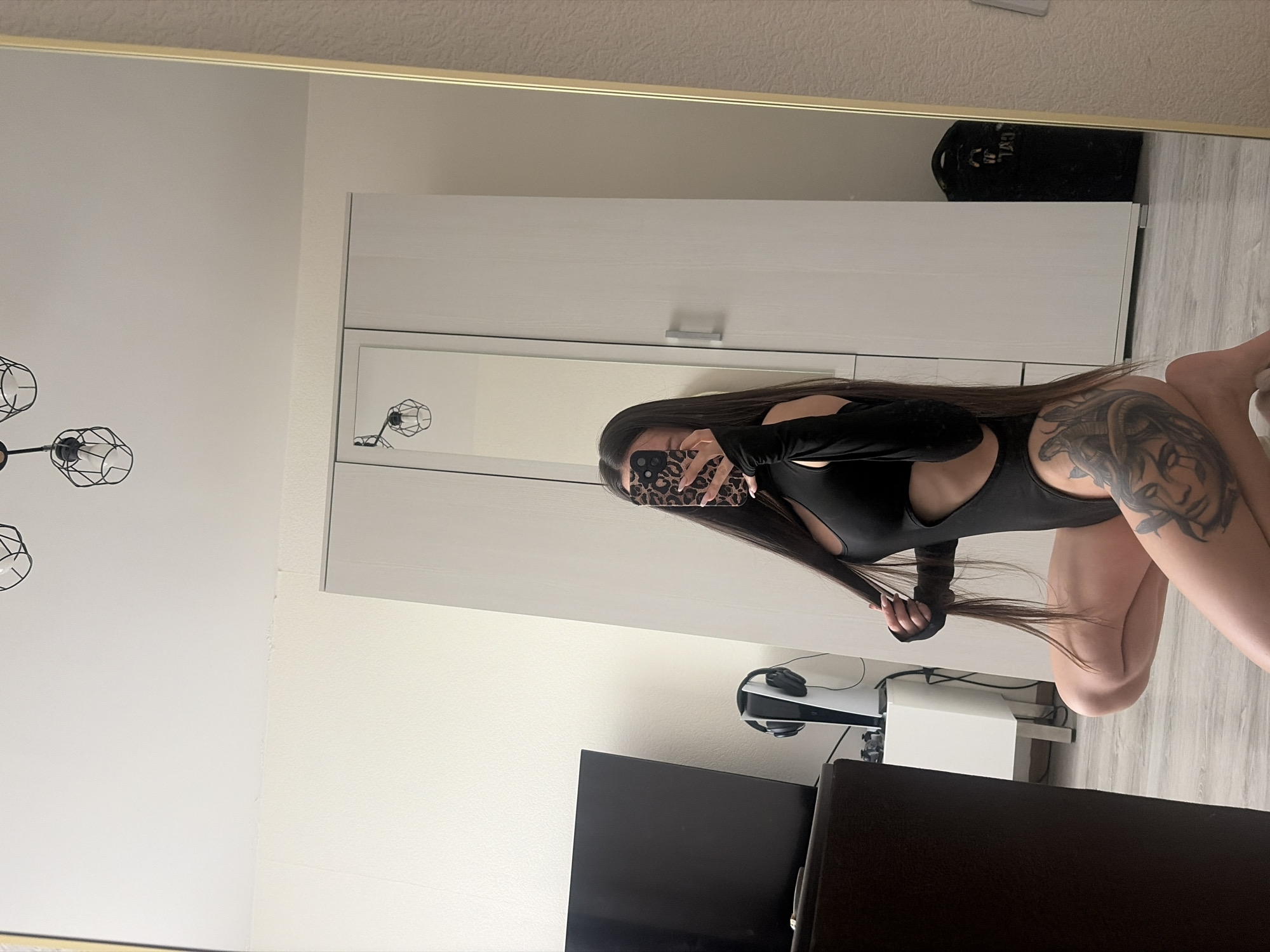 Here is a forensic description of the image:
The image shows a woman in a black bodysuit, posing in front of a mirror. She is holding her hair in her hand and her phone covers her face. Her right thigh is visible, and a tattoo is present on it. Behind her, there's a white closet or wardrobe with silver handles. Below the closet is a white side table with a Playstation 5 console on it. A black TV screen is placed in the foreground, partially blocking the view. The background is a plain wall. The location is an indoor setting, but the specific city and state are unknown.