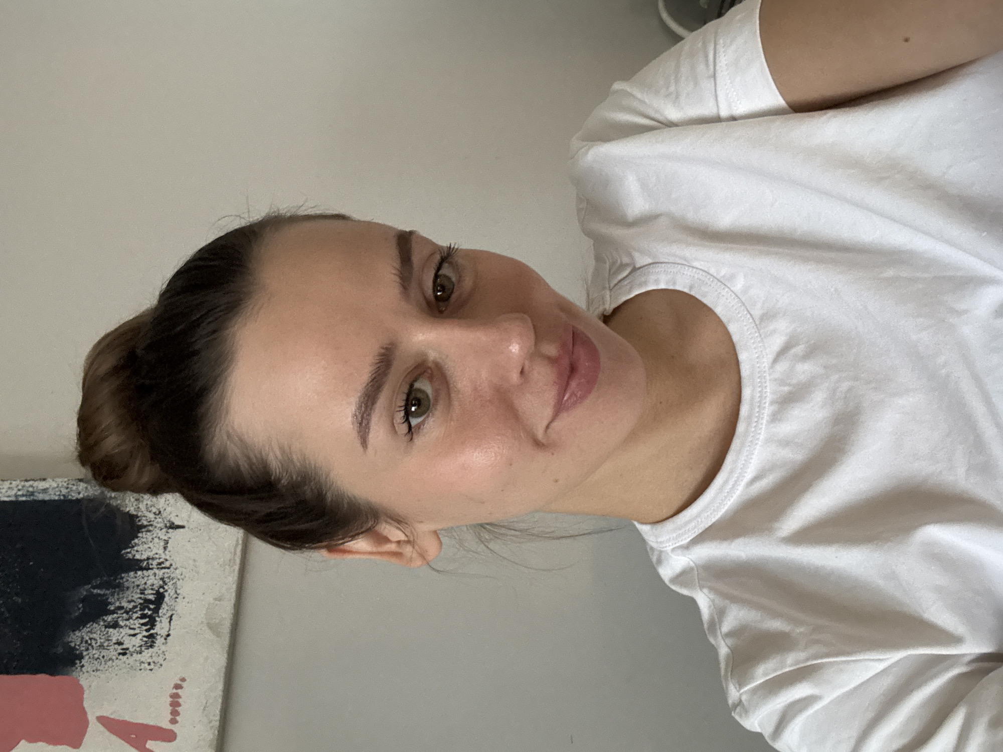 A fair-skinned woman with brown hair pulled back into a bun and green eyes is looking directly at the camera with a slight smile. She is wearing a plain white t-shirt. The background consists of a pale gray wall and a piece of abstract art in the bottom left corner featuring black, white, and pink paint. There is no indication of the location being in Krasnoyarsk, Russia.