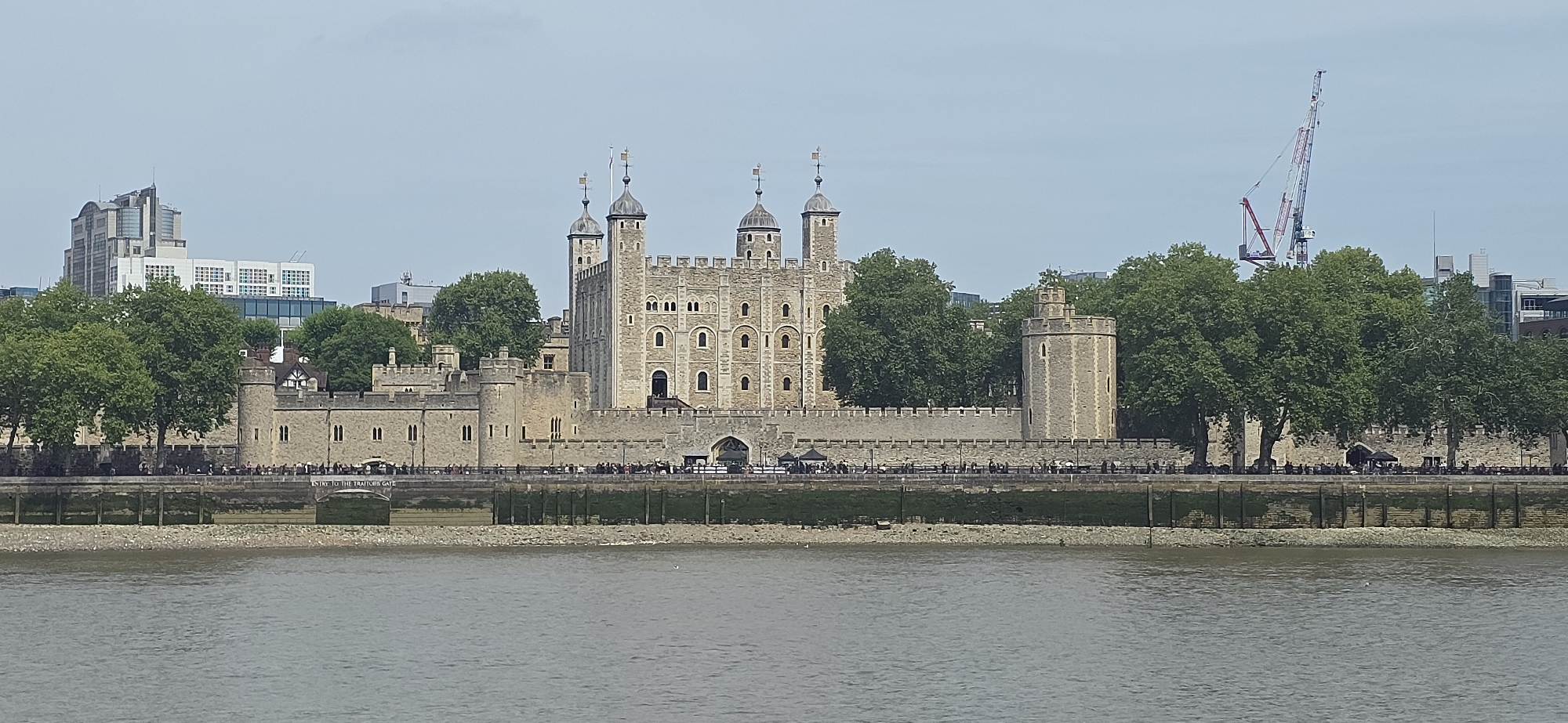 The image depicts the Tower of London, a historic castle located in central London, United Kingdom, viewed from across the River Thames. The Tower's White Tower is prominent, surrounded by other towers and fortifications. Trees partially obscure the base of the tower complex. A construction crane is visible behind the trees on the right side of the frame. Buildings are visible in the background to the left of the tower. The Thames is in the foreground, with a visible shoreline in between the river and the castle. People are gathered along the walkway in front of the castle.