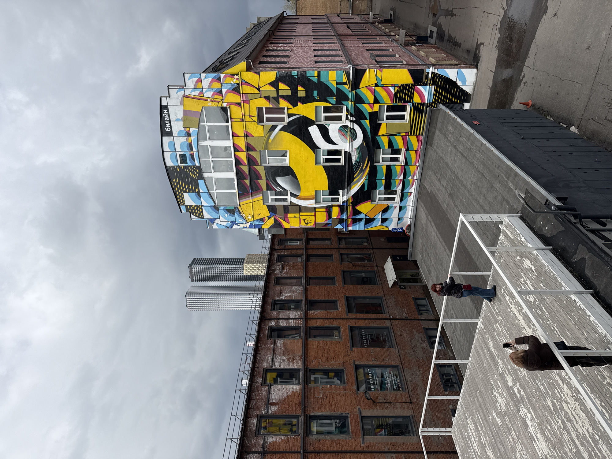The image captures a vibrant urban scene in Moscow, Russia, under an overcast sky.
A multi-story building dominates the right side of the frame, completely covered in an elaborate, abstract graffiti mural. The mural is a riot of color, primarily yellow, black, blue, pink, and white, featuring geometric patterns, wave-like motifs, and striped designs. A large yellow circle with a prominent stylized white number '6' or similar symbol is a central element of the artwork. The word "билайн" (Beeline), a Russian telecommunications company, is clearly visible in white Cyrillic script on a black vertical panel on the building's upper left side. The building itself has numerous rectangular windows integrated into the mural.
To the left of the graffitied structure stands an older, multi-story brick building with many dark rectangular windows. Some windows display signs with Russian text, including "КОНСТРУКТ" (KONSTRUKT). Further in the background, towering modern skyscrapers with reflective facades rise against the cloudy backdrop, contrasting sharply with the older architecture.
In the foreground, on a raised wooden deck with white railings, two individuals are present. One person, with reddish hair, stands facing the viewer, dressed in a dark jacket, blue jeans, and carrying a red shoulder bag. The second person, partially visible and facing away, is holding a smartphone, seemingly capturing a photo or video of the scene.
The ground level to the far right shows a cracked asphalt or concrete surface with a small orange traffic cone. The weather appears to be overcast daytime.