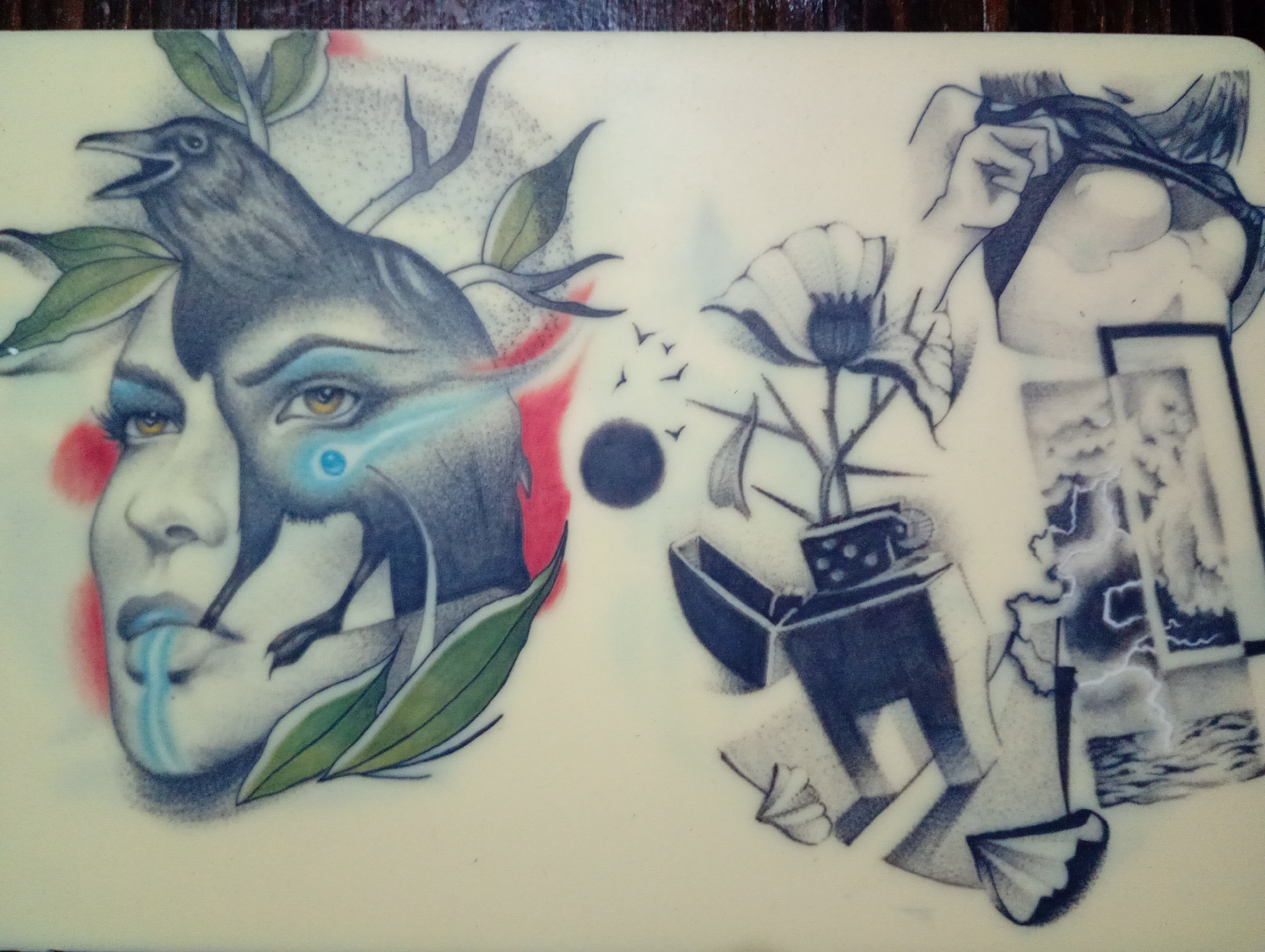 The image depicts a rectangular surface with two distinct tattoo designs.
On the left, a woman's face is partially obscured by a dark crow, with its legs appearing as if they are emerging from her face. She has light skin, bright gold eyes, and blue eyeshadow. Her lips are a shade of blue, with a blue stream emanating from her mouth. Green leaves are present near her face and the crow's body.
On the right, the design features a lighter with a flower growing out of it. A woman's hand is visible, and there is a framed picture showing a storm. A small dark circle is in the middle of the surface, and small bird silhouettes are flying away from it.
There is no indication that this image takes place in Roza, Bulgaria.