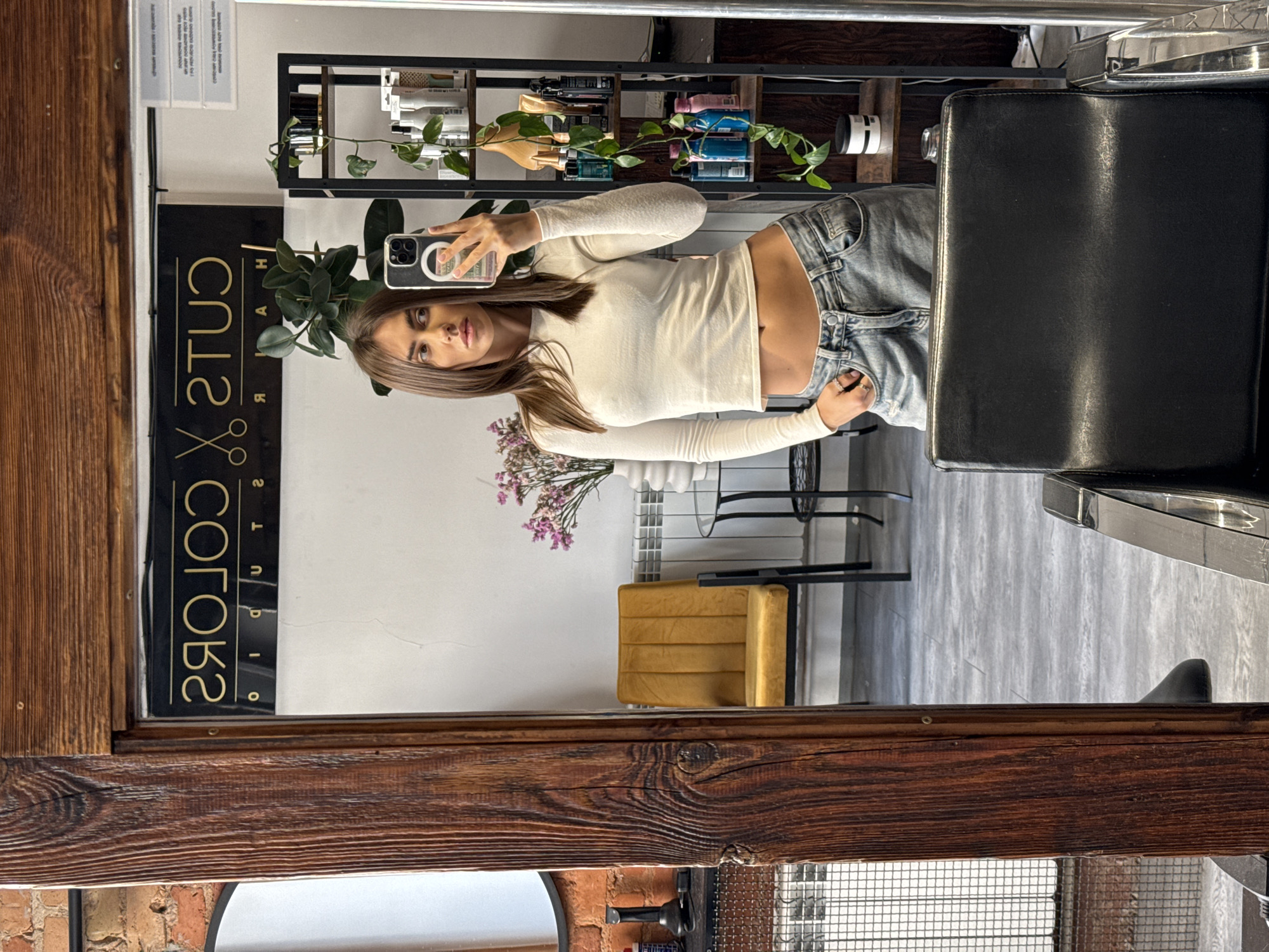 A woman with long brown hair is taking a selfie in a mirror, reflecting a salon or hair studio environment. She's wearing a long-sleeved white top and jeans. Behind her is a salon setting, including a black chair, shelving stocked with hair products, and decorative plants. A sign reading "CUTS COLORS STUDIO" is visible in the reflection. The location appears to be a professional hair salon in Warsaw, Poland, based on the salon's name and typical furnishings.