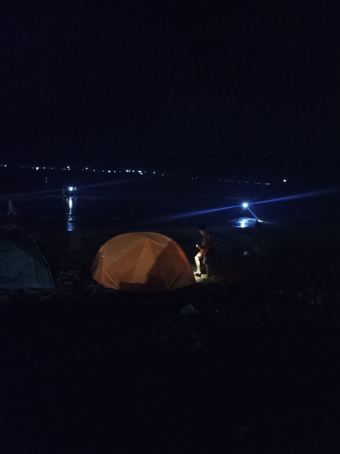 The image depicts a serene nighttime scene of camping outdoors in Kecamatan Pujut, Indonesia. In the foreground, an orange dome tent is prominently featured on the right side, appearing illuminated, possibly from within or by a nearby light source. To its left, a darker, possibly green or blue, tent is partially visible.
A person is standing near the orange tent, facing towards it, and appears to be holding a small, illuminated object, possibly a phone or a flashlight, which casts a soft glow around their feet. The immediate surroundings are dark, suggesting an open, possibly sandy or earthy terrain.
In the mid-ground and background, a large body of water is implied, reflecting a brilliant, elongated light source in the distance, which could be a boat or a light from the shore. The horizon is dotted with numerous small, bright lights, indicating a distant coastline, village, or other human activity. The overall ambiance is dark and tranquil, characteristic of a night spent camping by the water. There is no visible text in the image.