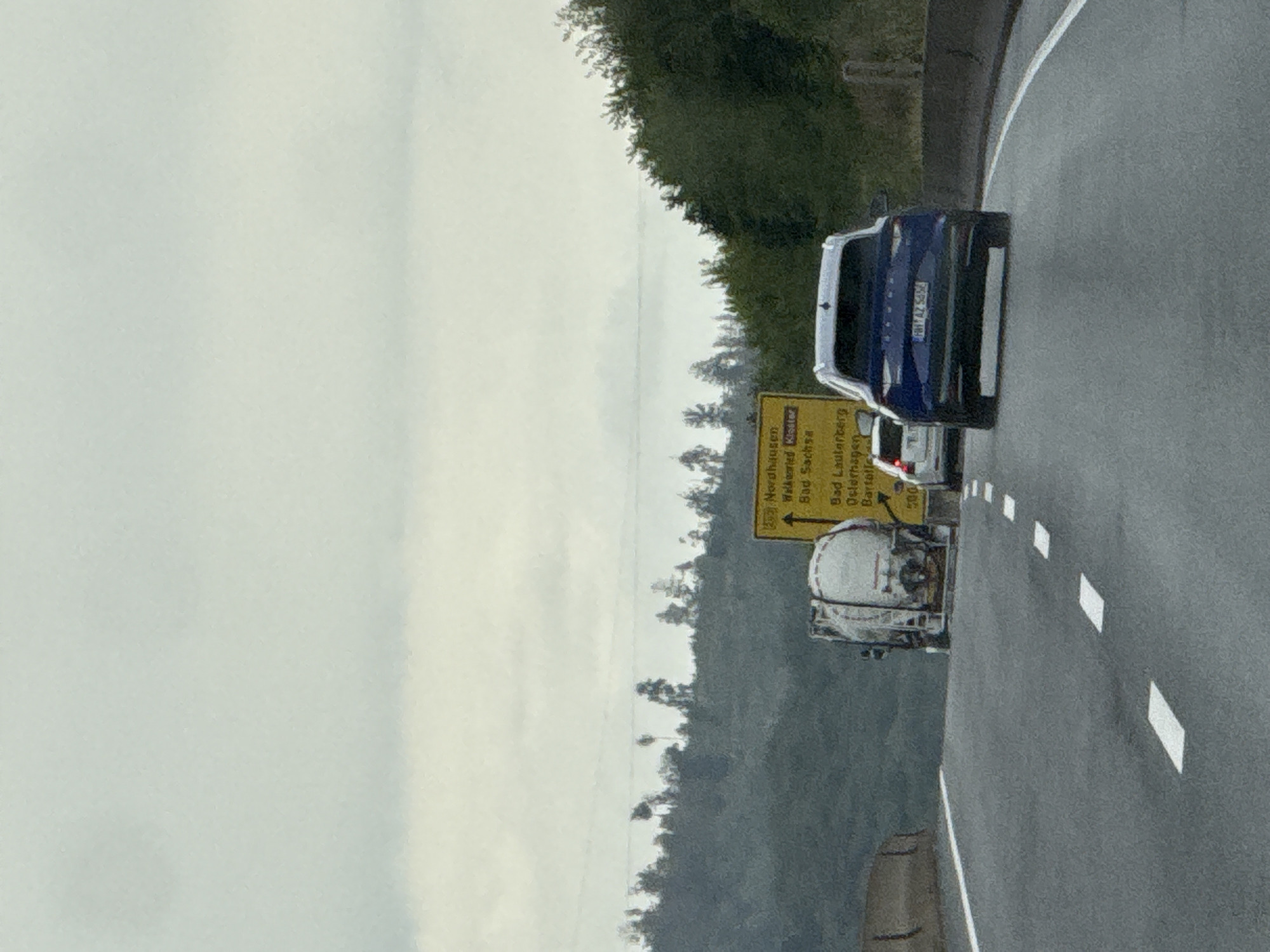 The image depicts a highway scene on a cloudy day, likely within or near Bad Lauterberg im Harz, Germany. In the foreground, a grey asphalt highway with white lane markings is visible. A dark blue SUV is driving in the right lane, with a white license plate that reads "FH-AZ 5856". Ahead of the SUV and slightly to the left is a white tanker truck, also on the highway. To the left of the tanker truck, a concrete barrier is visible. In the background, a yellow road sign with German text indicating directions to Nordhausen, Walkenried Kloster, Bad Sachsa, and Bad Lauterberg im Harz is partially obscured by the tanker truck. The sign also shows distances, including "500". A line of trees and hills forms the backdrop of the scene, with a hazy sky overhead.