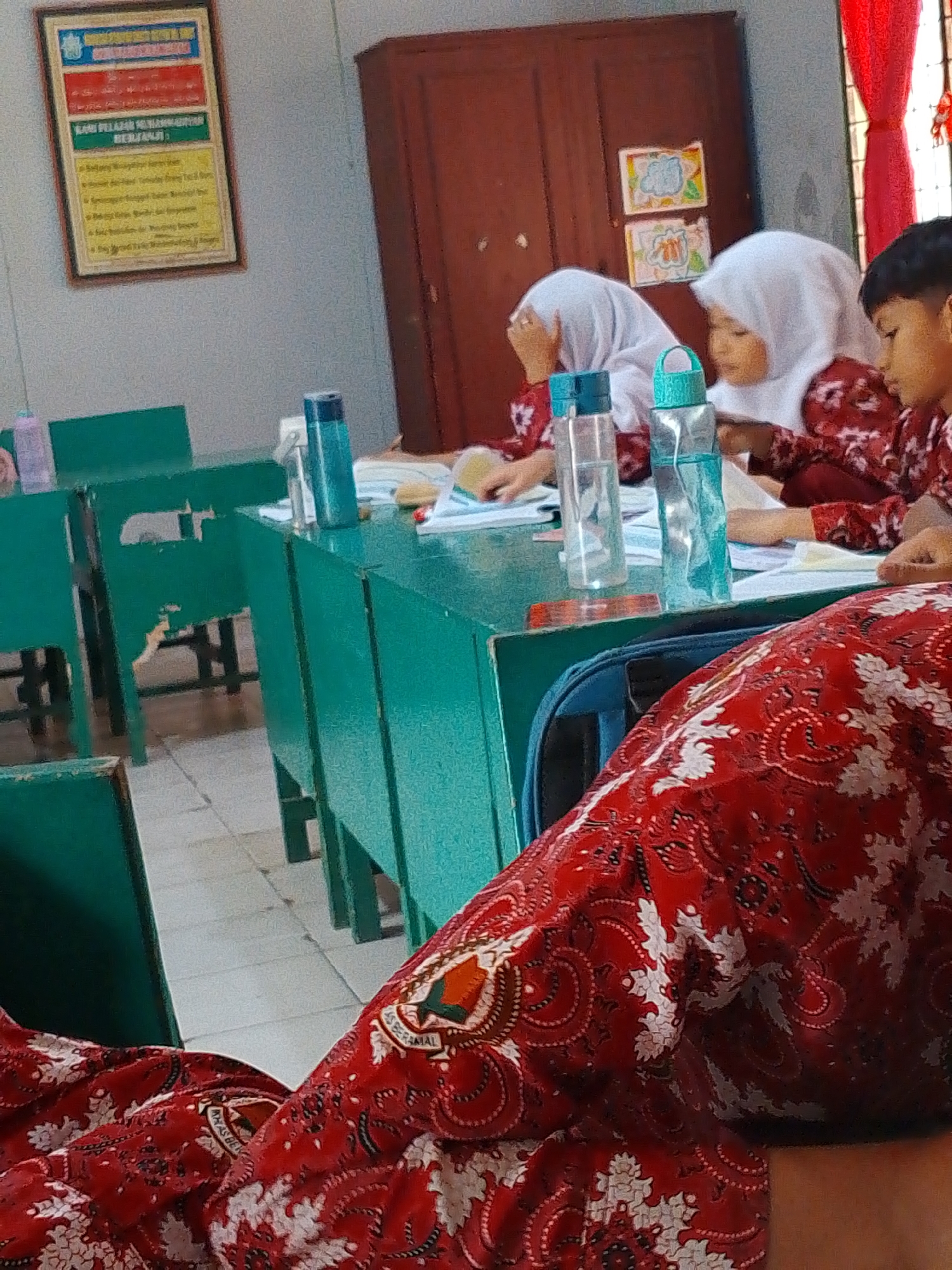 The image captures a classroom scene in Tembung, Indonesia, focusing on several students. In the foreground, a person wearing a red and white patterned garment is partially visible. Further back, two girls in white hijabs are seated at a teal-colored desk, appearing to be engaged in an activity, possibly reading or writing, with papers and water bottles on the desk. A boy, also wearing a red and white patterned garment, is seated to the right, looking towards the left. The classroom environment includes teal-colored desks and chairs, a wooden cabinet, and a framed poster on the wall displaying text in Indonesian. The lighting suggests an indoor setting, possibly during the daytime. The overall impression is that of a typical school day in Indonesia.