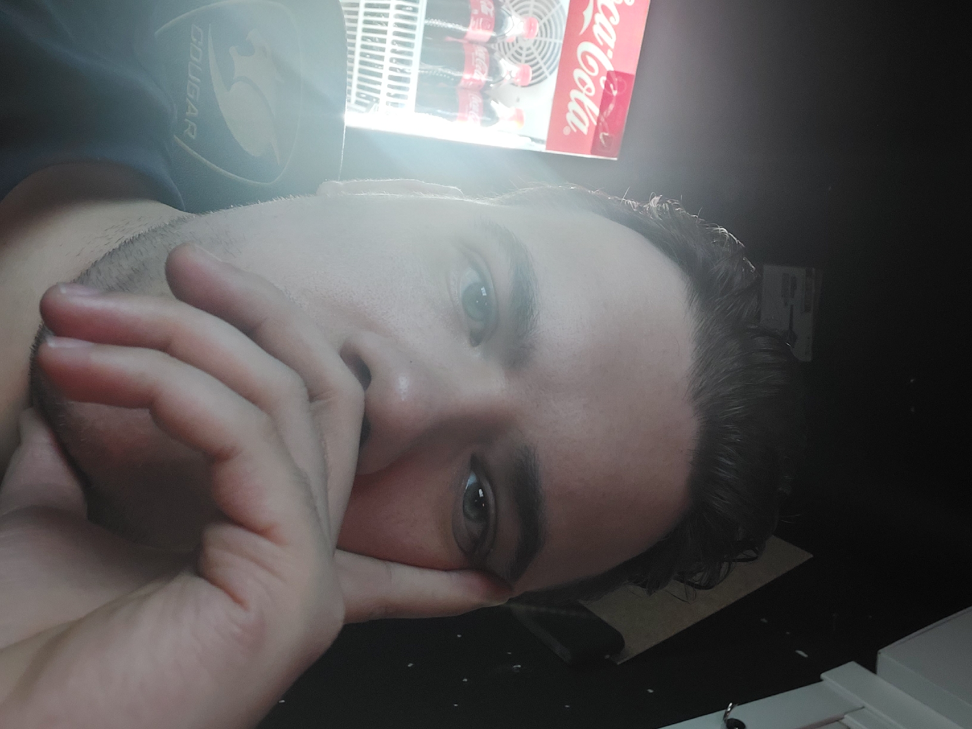 A man lies on his side, his face close to the camera. His hand is covering part of his face. The man has light skin, short dark hair, and light-colored eyes. He wears a navy blue shirt with a "Cougar" logo on the shoulder. A Coca-Cola vending machine stands behind him, with a bright light shining from the top. The location is unknown, and there is no basis to suggest it is in Tashkent, Uzbekistan.