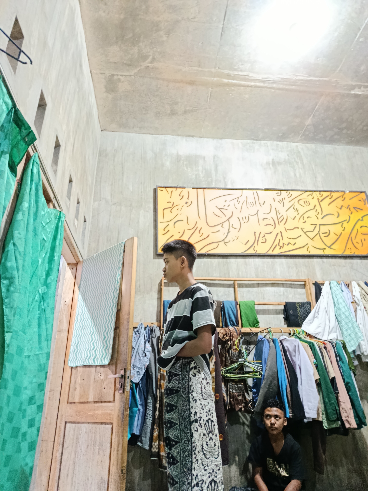 The image shows an indoor scene in Ramban Wetan, Indonesia. Two young males are present. The male in the foreground is standing, facing away from the camera, and appears to be looking towards a doorway. He is wearing a striped t-shirt and a patterned sarong. To his right, a second male is seated on the floor, facing forward and looking directly at the camera. He is wearing a dark t-shirt. The room has concrete walls and ceiling. A clothes rack filled with various garments is positioned against the wall behind the seated male. Green curtains hang to the left of the doorway, and a wooden door is ajar. A large, abstract, yellow artwork with Arabic calligraphy is mounted on the wall above the clothes rack. The environment appears to be a home or a storage area.