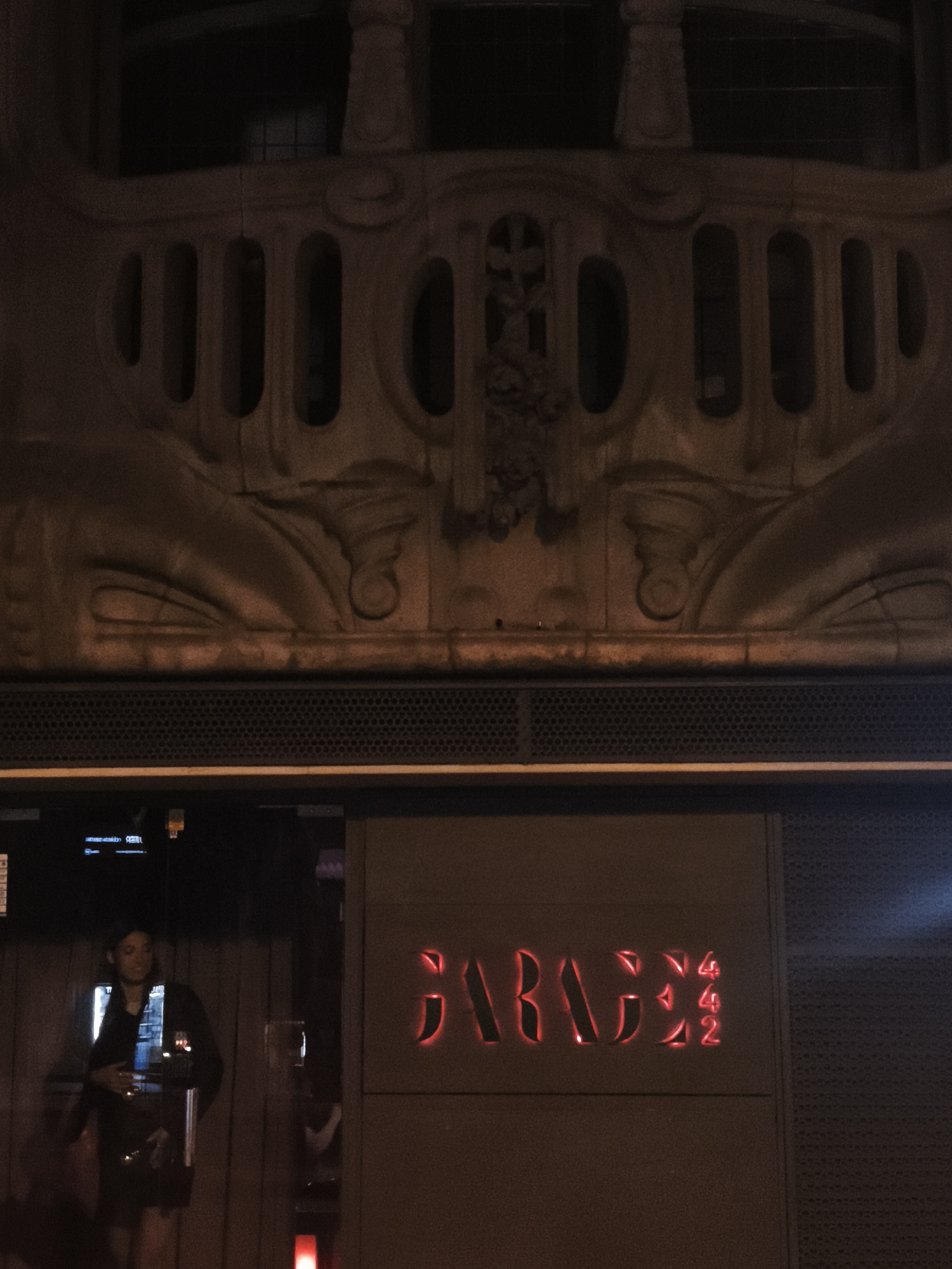 The image depicts the exterior of a venue at night. Above the entrance, an ornate, aged architectural facade with carved decorative elements is visible. Below this, a dark, perforated metal band runs horizontally across the image. The entrance itself is framed by a dark doorway and a sign with illuminated red lettering that reads "BARBEX 442". To the left of the sign, a woman in dark clothing stands partially obscured by a glass door. She appears to be an employee or doorman, holding what looks like a device or a purse. The lighting is dim, with the red sign being the most prominent source of light within the entrance area. The overall impression is that of a nightclub or bar in a somewhat aged urban setting.