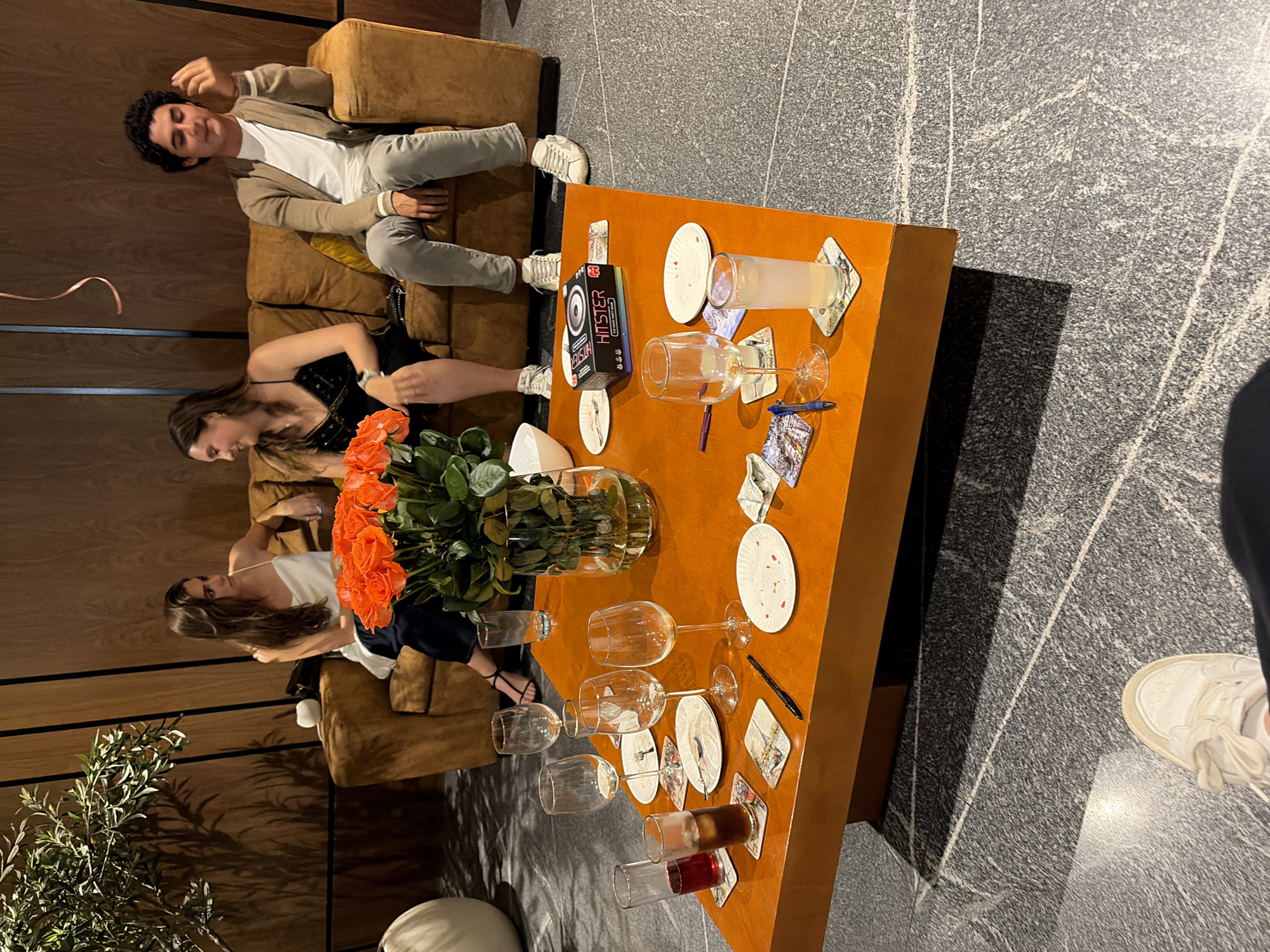 A high-angle, indoor shot depicts three individuals and a coffee table littered with plates, glasses, and other miscellaneous objects. A man sits on a brown couch, wearing a light tan jacket, white shirt, and light-colored pants with white sneakers. A woman in a black dress sits next to him on the same couch. Another woman, wearing a white top and dark bottoms, is seated on a matching brown couch to the left, partially obscured by an orange rose bouquet in a glass vase on the table. The coffee table, which is orange, is covered with an assortment of items including glasses, plates, playing cards, coasters, pens, and a game box labeled "Hister." The floor is grey with white lines. A potted plant stands in the background on the left. The location appears to be a lounge or living room. The image does not provide information about the location in the city of San Bernardino Tlaxcalancingo, Mexico.