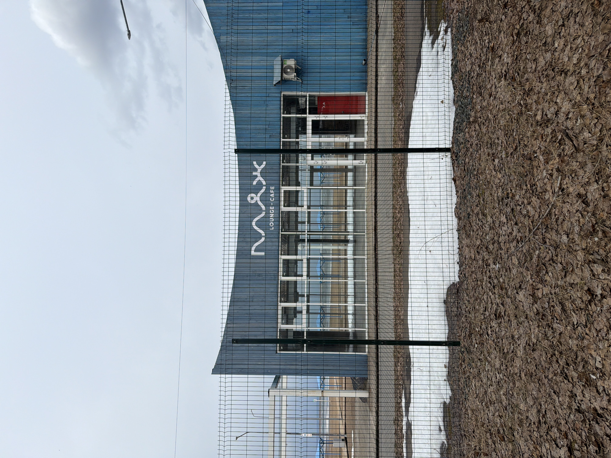 The image displays an outdoor scene featuring a blue building, a metal fence, and ground partially covered in snow and fallen leaves, under an overcast sky. The photograph is oriented vertically, showing the building on the left, the sky on the top, and the ground on the right when viewed upright.
The main subject is a modern, blue-paneled building with large windows. An external air conditioning unit is visible on the upper part of the building's facade. A prominent white sign on the building features a stylized logo that appears to read "NYX" or a similar character set, with the text "LOUNGE - CAFE" written vertically below it. One windowpane on the building's upper floor reflects a red color, possibly from an interior curtain or object.
In the foreground, a dark green chain-link or mesh fence runs horizontally across the frame, separating the viewer from the building and the ground beyond. The ground behind the fence is a mix of dried, brown leaves and patches of white snow, suggesting late winter or early spring weather conditions in Chaikovsky, Russia. The sky, visible above and to the left of the building (when viewed upright), is light grey and cloudy. No people or discernible activities are present in the static scene.
