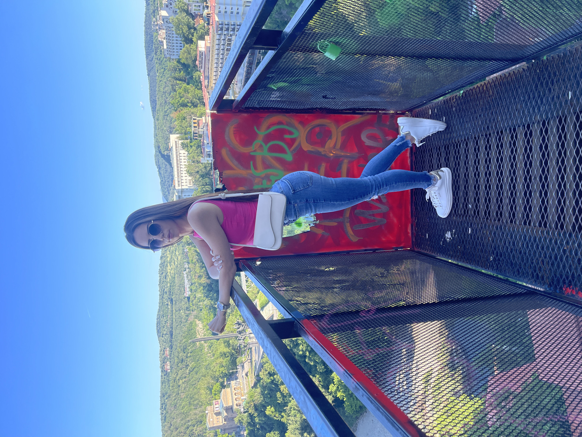 A woman is posing on a metal platform that appears to be an observation deck, overlooking a city nestled in a valley. She is wearing a pink tank top, blue jeans, white sneakers, and sunglasses. A white crossbody bag is slung over her shoulder. The platform has a black metal railing and a red panel on the right side adorned with graffiti. A green padlock is attached to the railing. In the background, buildings are visible amidst lush green hills under a clear blue sky. The platform's floor is made of black metal grating.