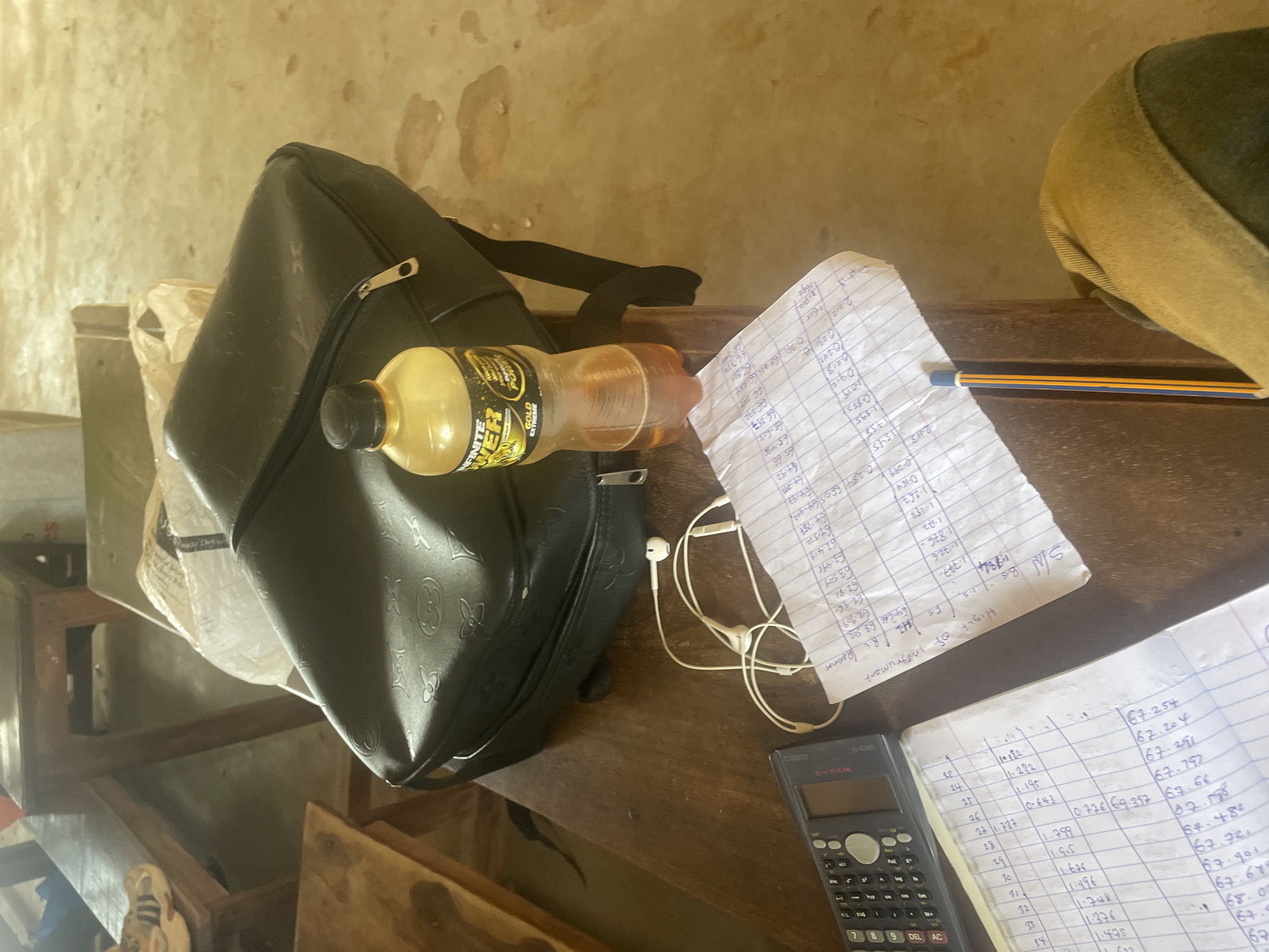 The image depicts a desk in what appears to be a classroom or study area in Lokoja, Nigeria. On the desk are a black backpack, a bottle of "Infinite Power" energy drink, a pair of white earphones, a calculator, and several sheets of paper filled with handwritten numbers and calculations. A yellow and blue pencil lies on top of one of the papers. The desk is made of dark wood, and the floor is a light brown, mottled surface. The background shows wooden benches and what looks like a wall. The lighting suggests it is daytime.