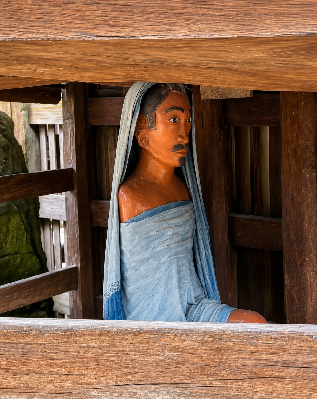 A carved wooden statue of a man, with orange skin and a blue cloth draped around him, is seated within a wooden structure. The man has a stern expression, with dark hair styled with a comb-like pattern, and a thin mustache and goatee. The statue is positioned under a wooden beam and in front of vertical wooden planks. To the left of the statue, a large, moss-covered rock is partially visible, along with a wooden fence. The overall scene appears to be an outdoor setting within the historical village of Ambarita, Indonesia, likely part of the Ambarita Stone Chairs site, which is known for its ancient Batak stone furniture and artifacts.