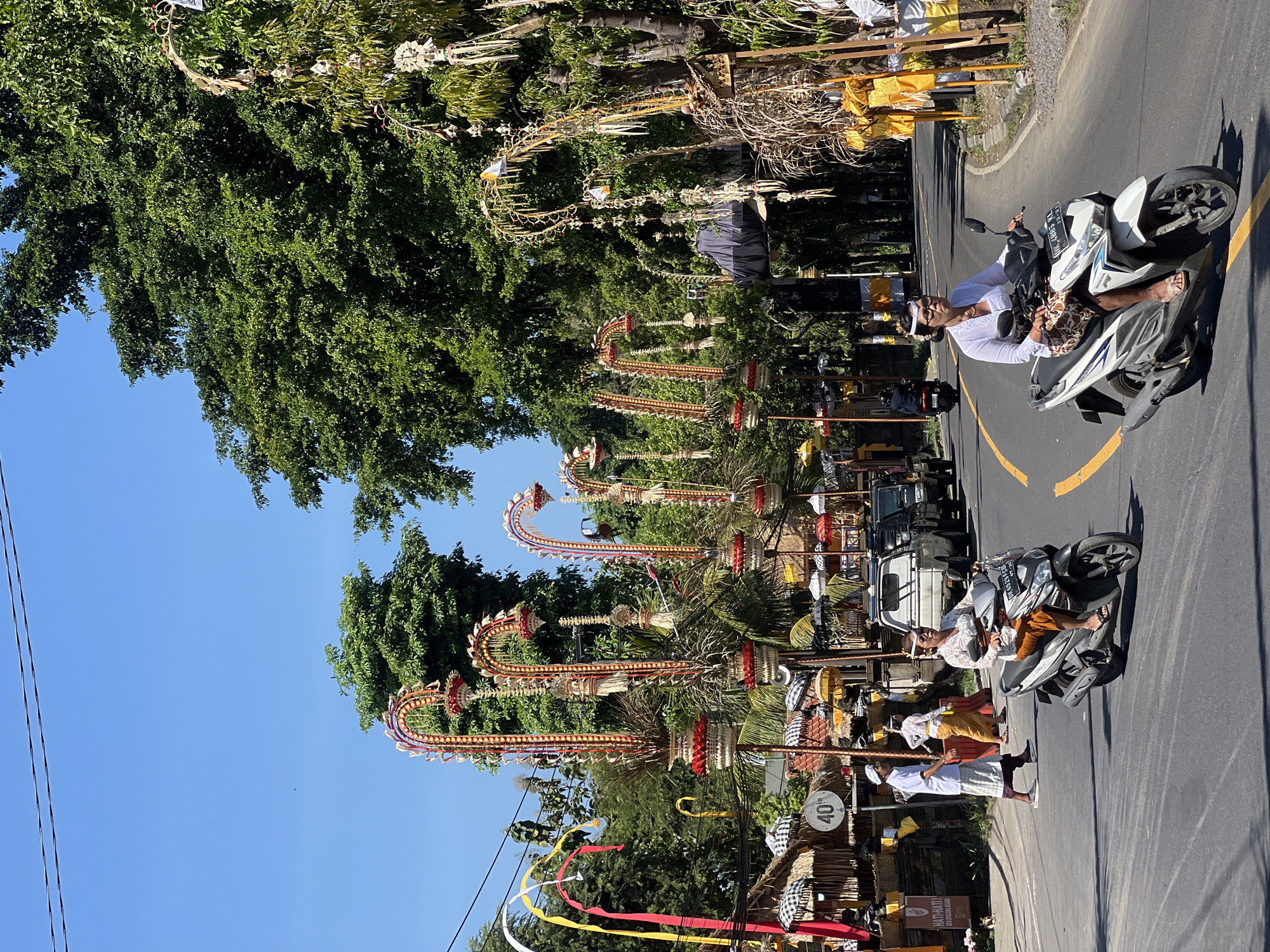 In Sukawati, Indonesia, a street is lined with elaborate, decorative arches, some adorned with yellow and red fabric streamers, others with woven palm fronds and white ornaments. Several people in traditional Balinese attire are visible. A woman wearing a white top and a patterned sarong rides a white scooter in the foreground. Behind her, a man in a white shirt and sarong walks on the sidewalk. Further down the road, another woman in similar traditional clothing rides a scooter. The street is paved with asphalt and bordered by lush green trees. Buildings with thatched roofs and patterned umbrellas line one side of the street, and several vehicles, including pickup trucks, are parked or driving on the road. A speed limit sign indicating "40" is visible on a pole. The sky is clear and blue, suggesting a sunny day.