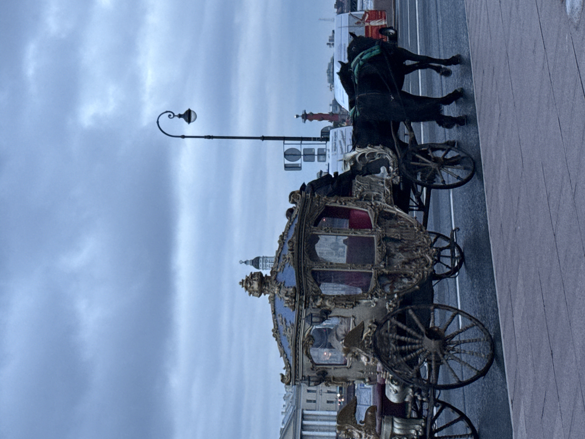 This is a medium shot photograph taken on a cloudy day, likely in Saint Petersburg, Russia. The foreground shows a paved pedestrian walkway. Behind it, on a wet asphalt road, a horse-drawn carriage is visible. The carriage is ornately decorated with gold trim and a blue roof, and appears to be carrying at least one passenger inside, with a driver visible at the front. Two black horses are harnessed to the carriage, with green harness elements. In the background, buildings with classical architecture, including columns, are present. A prominent street lamp with a curved arm and a light fixture stands to the right of the carriage. Further in the distance, a red obelisk-like structure and a white bus are visible. The overall mood of the image is somewhat somber due to the overcast sky and wet conditions.