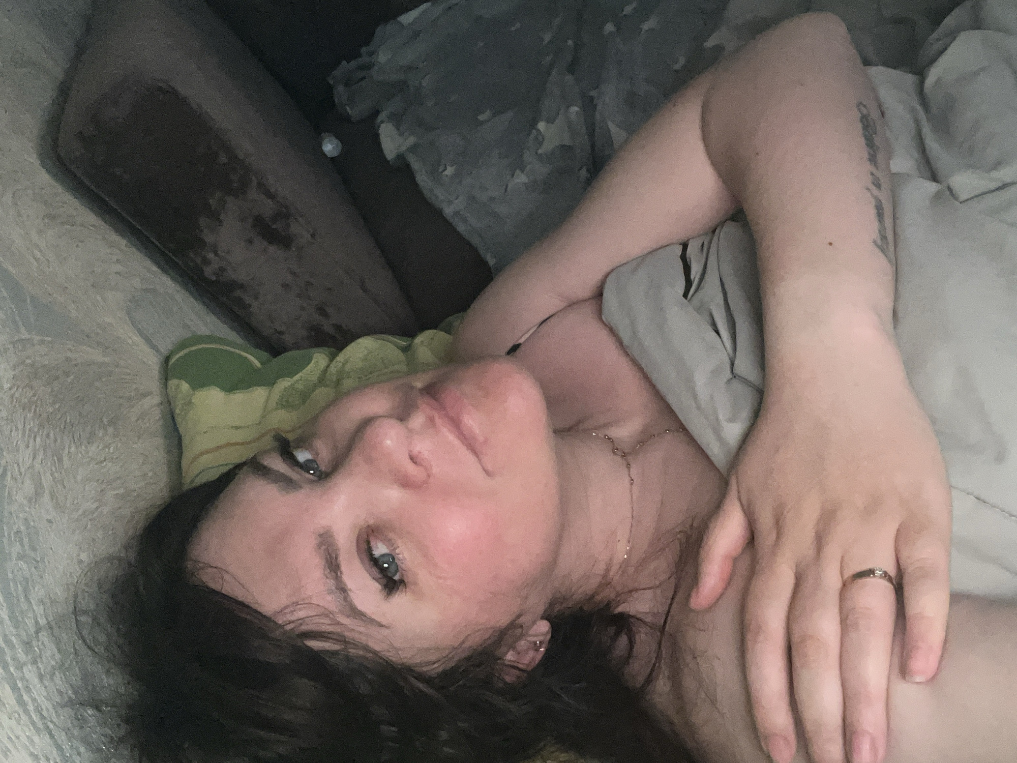 A fair-skinned woman with dark hair and blue eyes is lying on a bed. She is facing the camera, with her head resting on a yellow and green patterned pillow. A light gray blanket covers part of her body. She is wearing a delicate gold necklace and a ring on her ring finger. A tattoo is visible on her left arm. The background includes a dark brown object and a gray blanket with star patterns. No information indicates this photo was taken in Noviy, Russia.