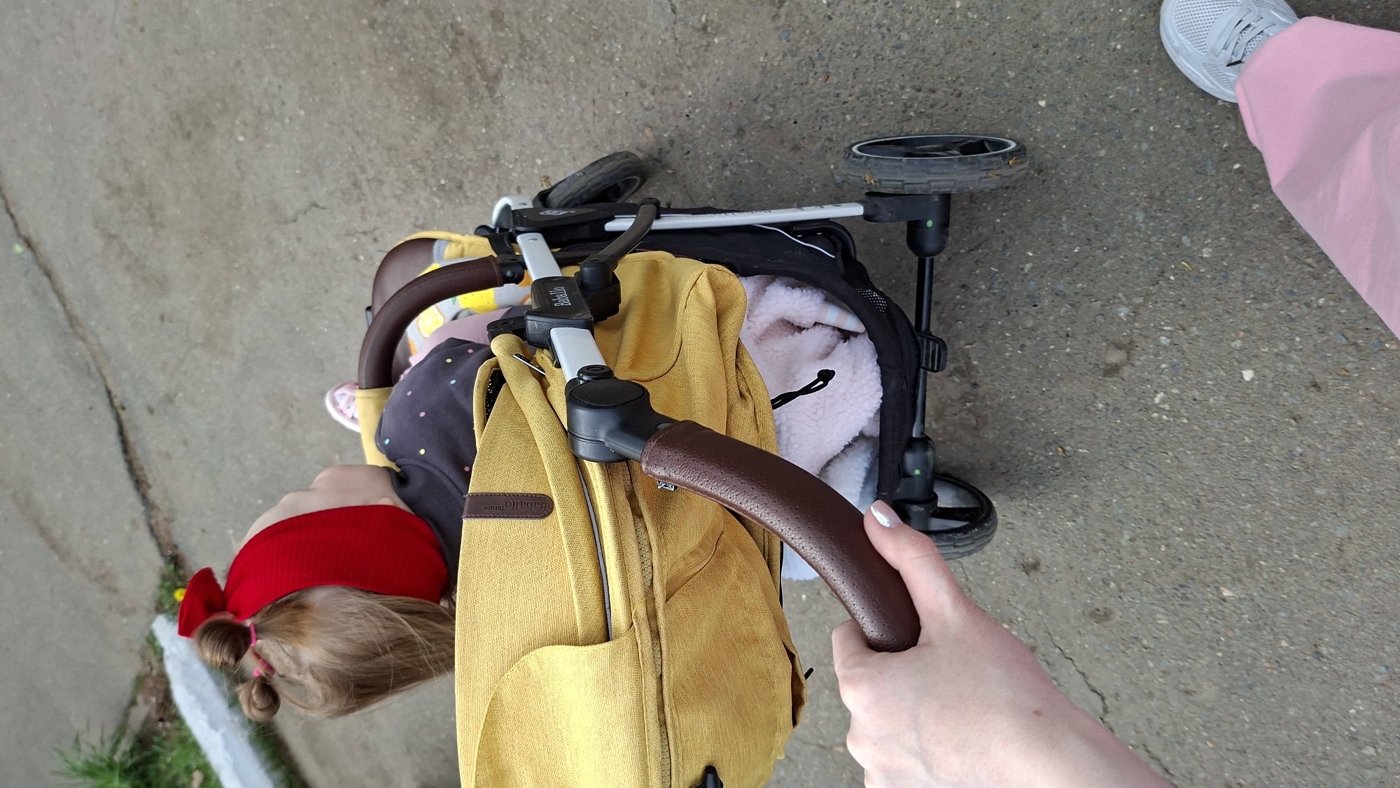 The image shows an overhead view of a baby stroller on a concrete surface. A child wearing a red headband with a bow is visible in the stroller. The stroller has a yellow diaper bag attached to the handle. A hand is holding the stroller handle. A foot wearing a gray sneaker and pink pants is partially visible at the top right of the image. The scene appears to be located outdoors on a sidewalk or pavement. Given the user's query includes "Chaikovsky, Russia", it is implied the image was taken there, though there are no explicitly visible indicators within the image confirming this location.