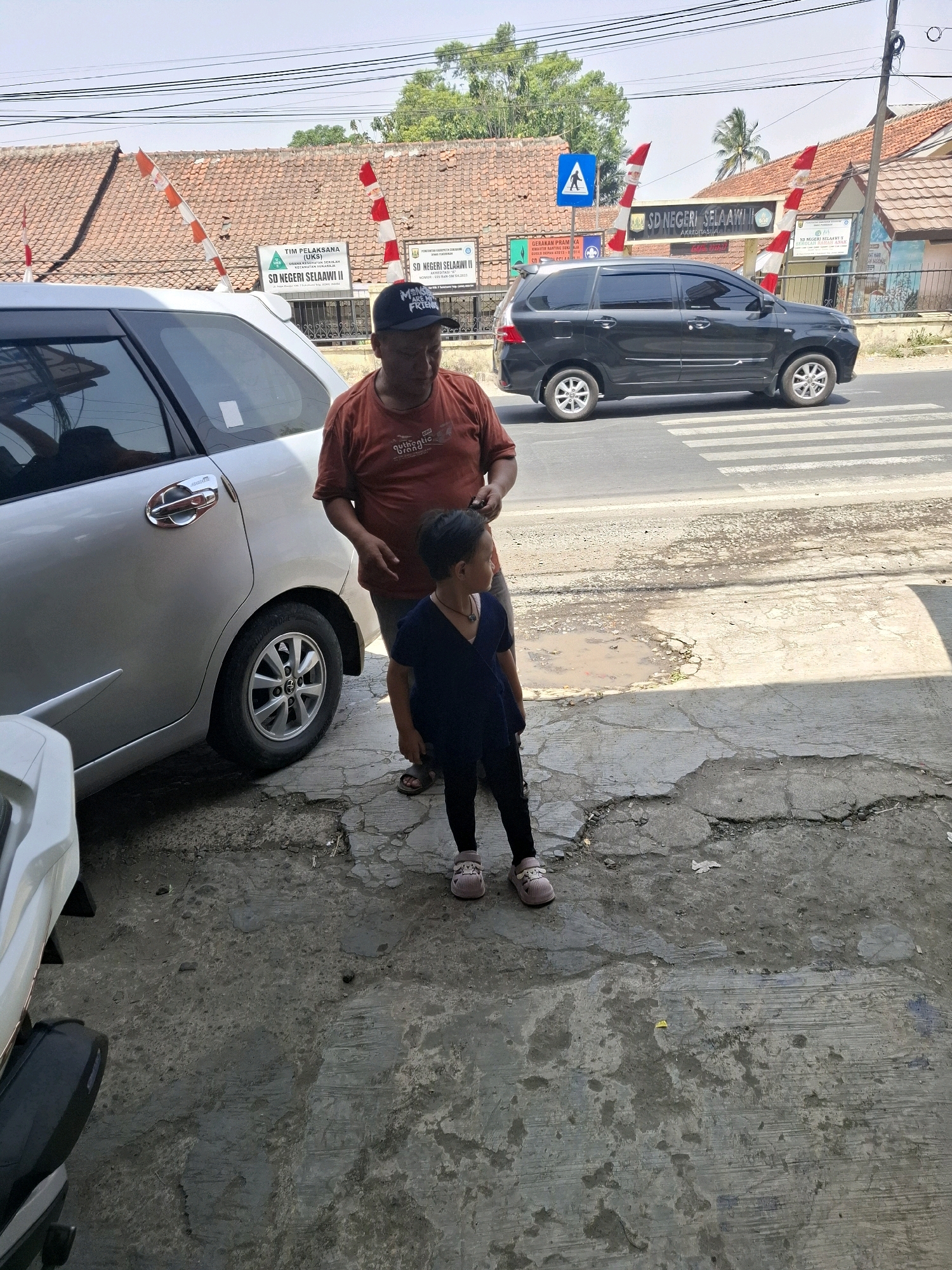 In Selaawi, Indonesia, a man and a young child stand on a cracked concrete surface in front of a silver vehicle. The man, wearing an orange t-shirt and a black baseball cap, faces away from the camera. The child, dressed in a dark blue top and black pants, looks to the right. Behind them, a black car drives on a road with a pedestrian crossing. A building with a red-tiled roof, displaying a sign for "SD NEGERI SELAAWII II," is visible in the background, along with Indonesian flags. The sky is clear and blue, suggesting it is daytime.