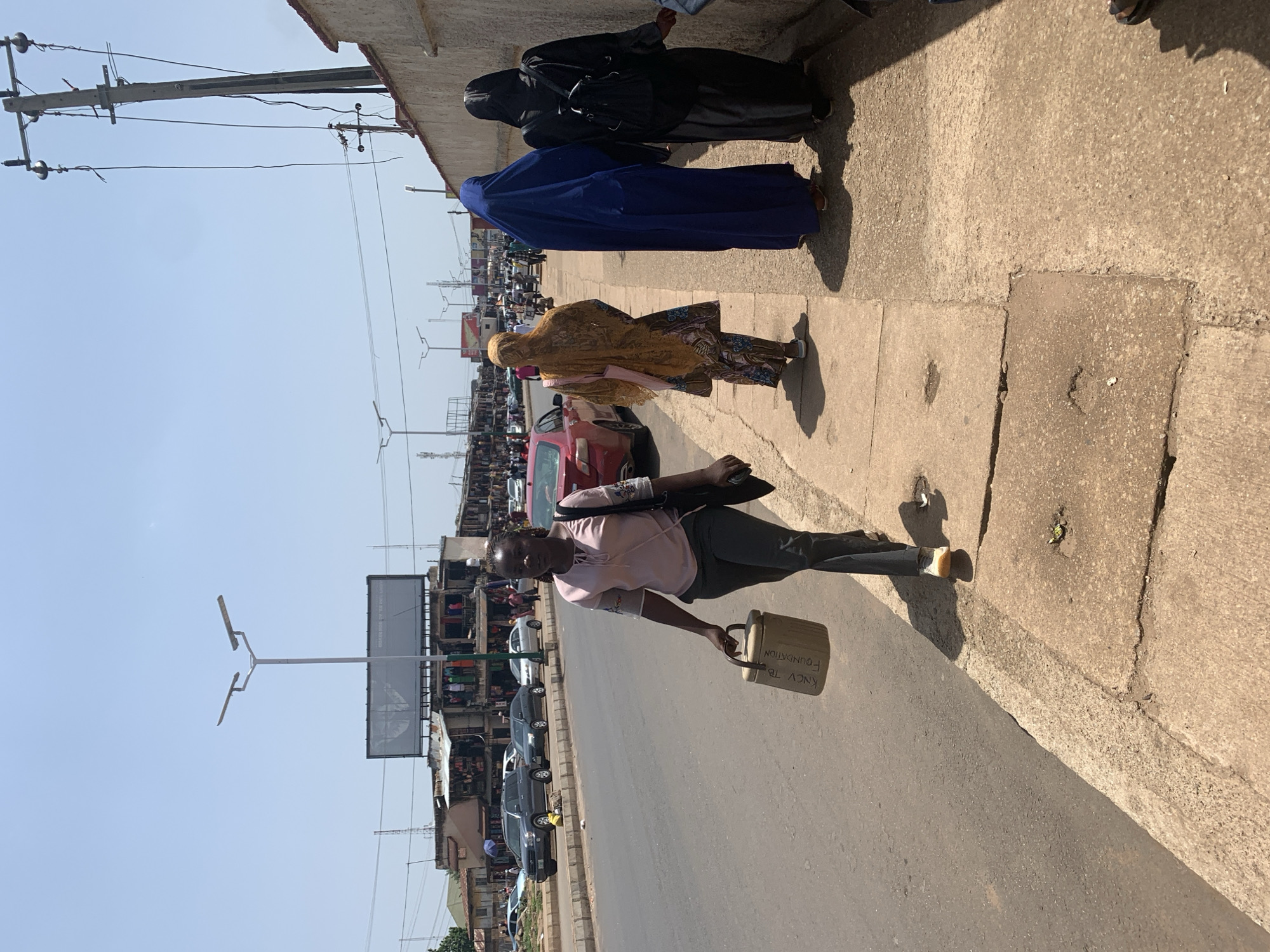 In the city of Jos, Nigeria, on a sunny day, four women are walking on a sidewalk next to a street lined with parked cars and buildings. A woman in the foreground, wearing a pink shirt and dark pants, carries a tan cylindrical container with writing on it. Behind her, three other women are walking away from the camera. The woman closest to the foreground is wearing a patterned yellow and orange outfit. The next woman is wearing a blue hijab, and the woman farthest back is wearing a black hijab and carrying a backpack. The street is paved, and the sidewalk is made of concrete. In the background, there are shops and market stalls, with people visible in them. Overhead, there are power lines and streetlights.