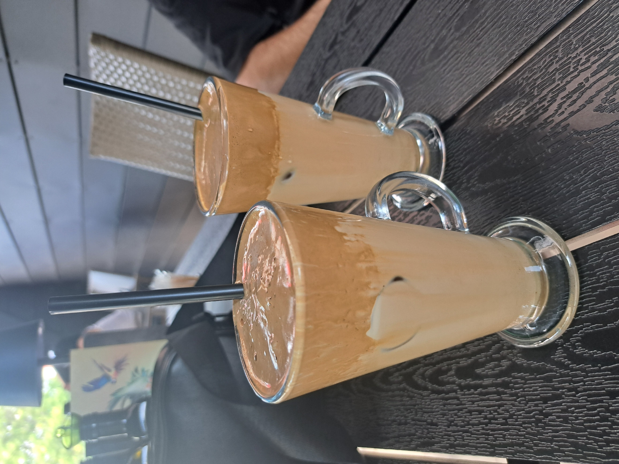 Two Dalgona coffees, each in a clear glass mug with a handle, sit on a dark wooden table with a textured finish. Both drinks have a layer of whipped coffee foam on top of a lighter liquid, with ice cubes visible in the front glass. Black straws extend from the top of each drink. In the background, a black bag is partially visible, along with a chair and a framed picture on a wall. The scene appears to be outdoors, possibly on a patio or deck, indicated by the presence of natural light and foliage in the distance.