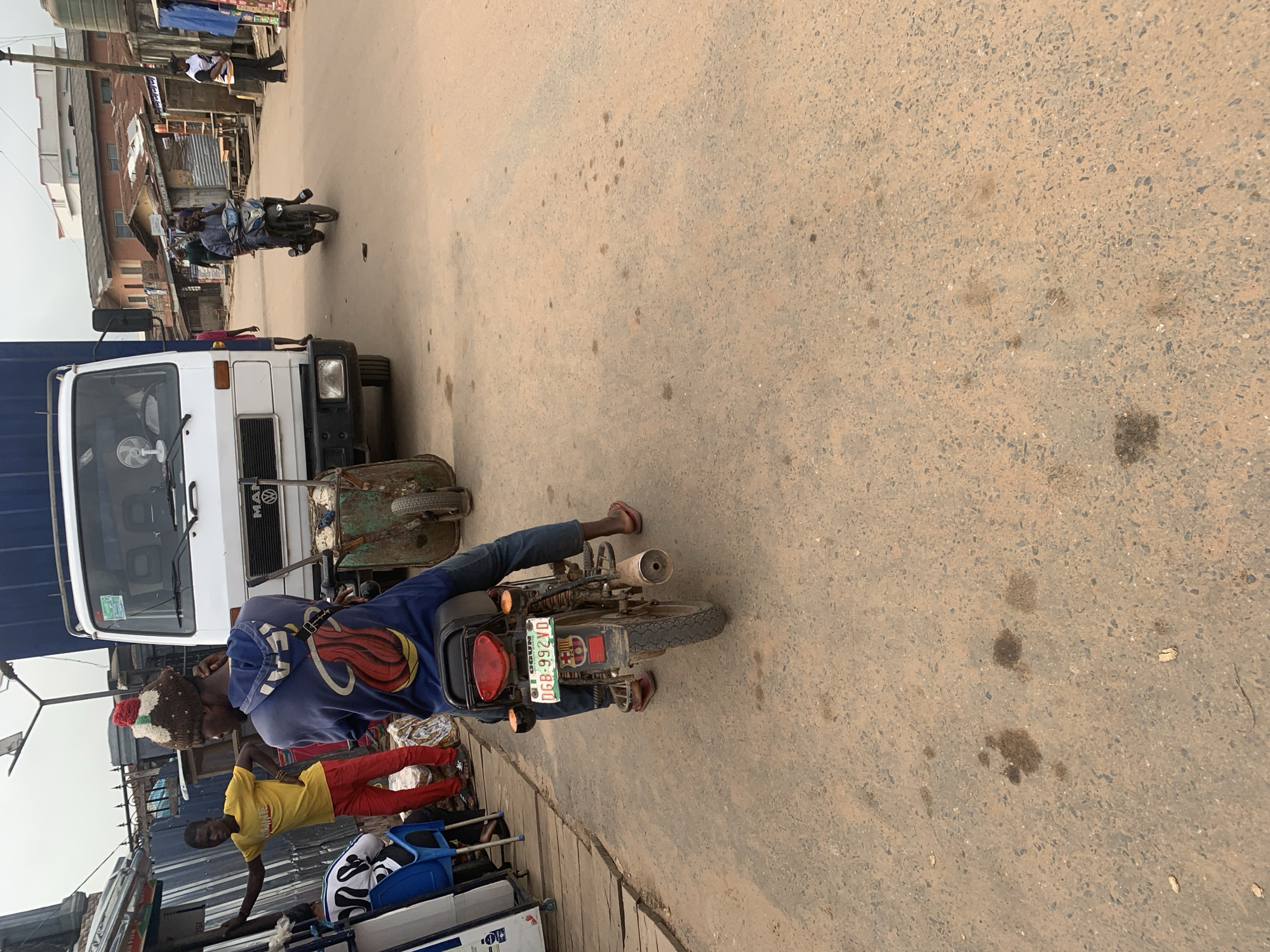 A medium shot captures a scene on a dusty, paved street in Shagamu, Nigeria, likely during daylight hours. In the foreground, a person wearing a blue hooded sweatshirt and red sandals is seated on a motorcycle. The motorcycle has a visible license plate that reads "OGUN DGB-992VD". Behind this motorcycle, a white truck is parked. To the left of the truck, another person wearing a yellow t-shirt and red pants stands, facing away from the camera.
The background reveals a bustling street environment with various buildings, some appearing to be made of corrugated metal and others of concrete. A few other individuals are visible in the distance, and there are also other vehicles, including what appears to be a three-wheeled vehicle or a small cart parked next to the truck. The overall impression is of a busy marketplace or a roadside area with ongoing activity. The ground is paved and shows signs of wear and tear, with some dark spots that could be oil or dirt stains. The sky is overcast, suggesting a cloudy day.