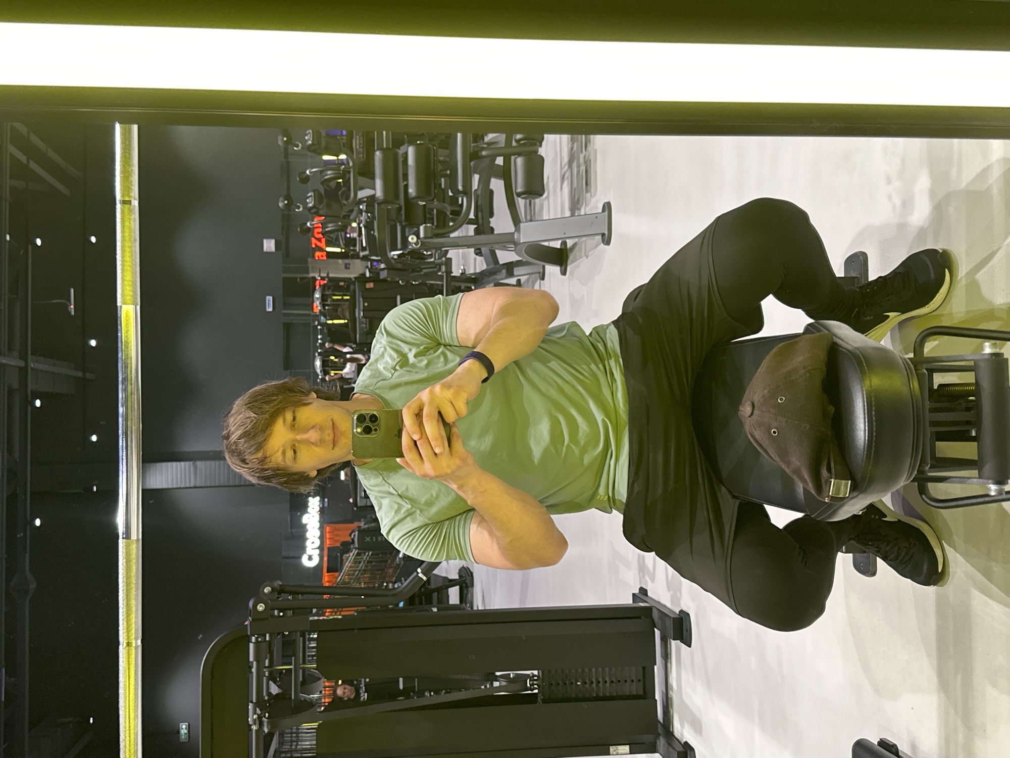 A person is taking a selfie in a gym. The person is wearing a light green t-shirt and black leggings. They are sitting on a weight machine with their legs extended. A brown baseball cap is on the seat of the machine. The gym appears to be well-equipped with various weight machines and other fitness equipment. The lighting in the gym is bright, suggesting it is daytime. There is a large mirror reflecting the scene. Visible text includes "CrossBox" on a sign in the background. The overall mood appears casual and focused on fitness.