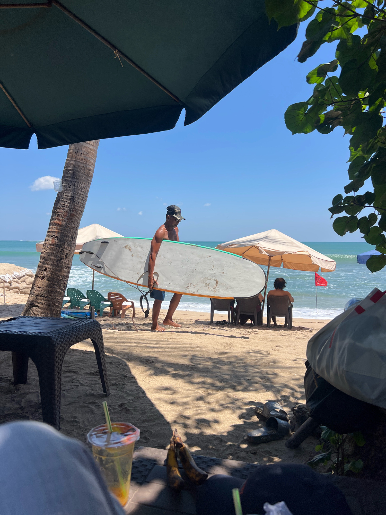 An outdoor scene on a sunny day at Kuta Beach, Indonesia. In the foreground, a person's legs are visible, with a drink and a pair of bananas resting on a surface. A dark rattan table is positioned to the left. In the mid-ground, a man walks across the sand carrying a surfboard. He is wearing a camouflage hat and shorts. Behind him, two beach umbrellas are set up, and several beach chairs are occupied by people. The ocean and waves are visible in the background under a clear blue sky. A palm tree stands to the left of the man with the surfboard.