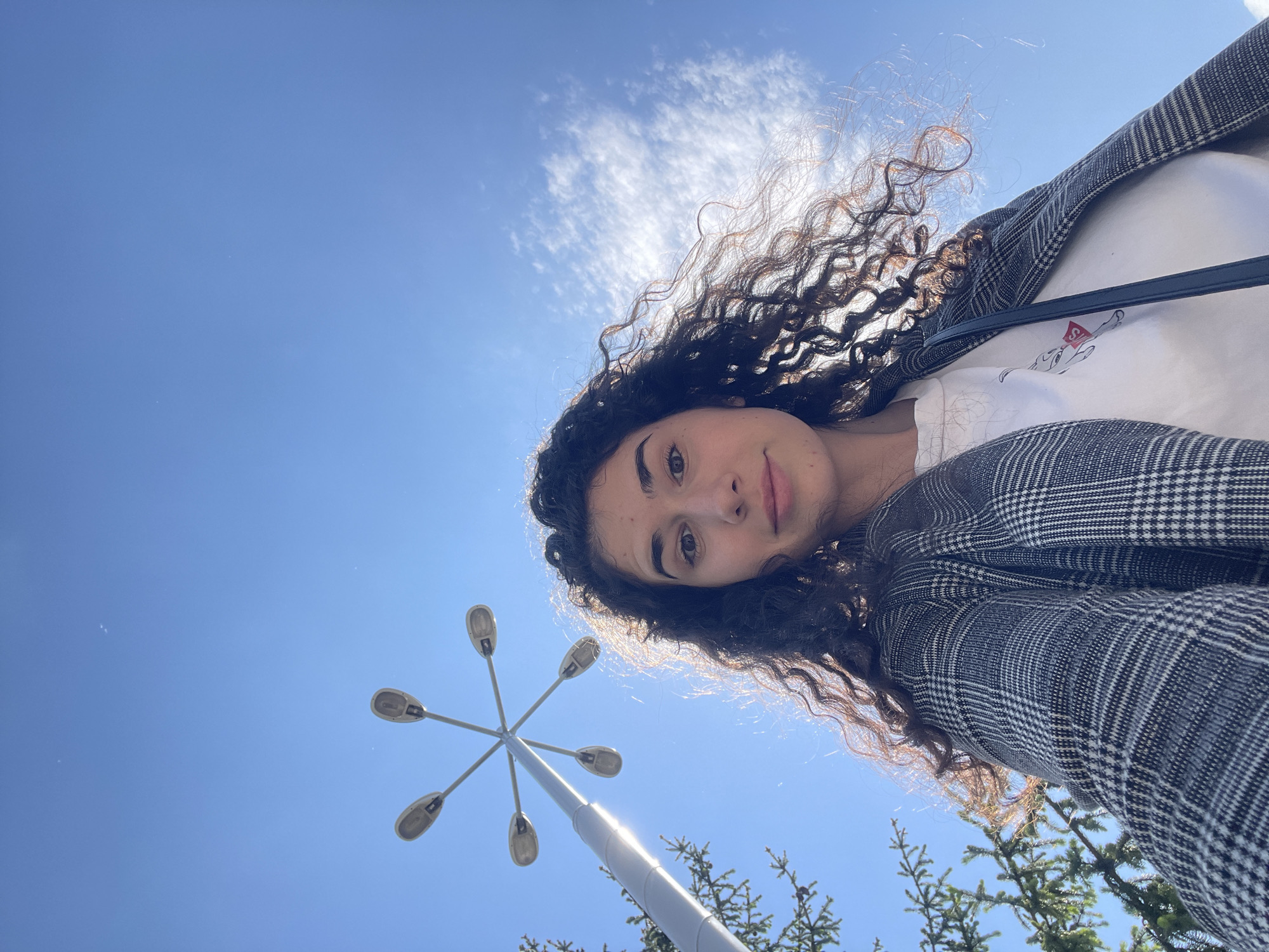 A low-angle selfie shows a young woman with curly brown hair. She is wearing a gray and white plaid jacket over a white shirt. A black purse strap is visible over her shoulder. Behind her is a bright blue sky with some wispy clouds. A streetlight with six lamps extends into the frame from the bottom left. Green trees are visible at the bottom right. The location is likely Sofia, Bulgaria, but this cannot be verified solely from the image's content.