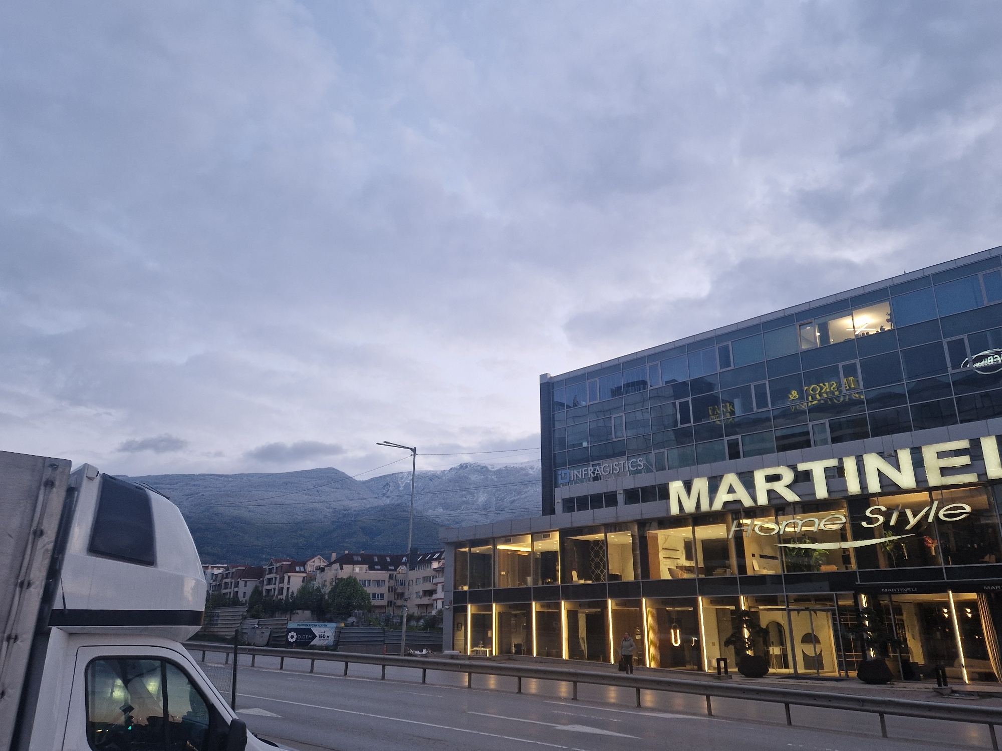 The image captures an urban scene on a cloudy day. A large building with glass windows and the sign "MARTINELLI Home Style" is prominently featured. Above it is another company called "Infragistics". The building is situated next to a road with a metal guardrail and a white van is visible on the left side. In the background, there are other buildings and a mountain range partially covered with snow. A person is walking along the sidewalk in front of the building. The location is in the city of Unknown, Unknown.