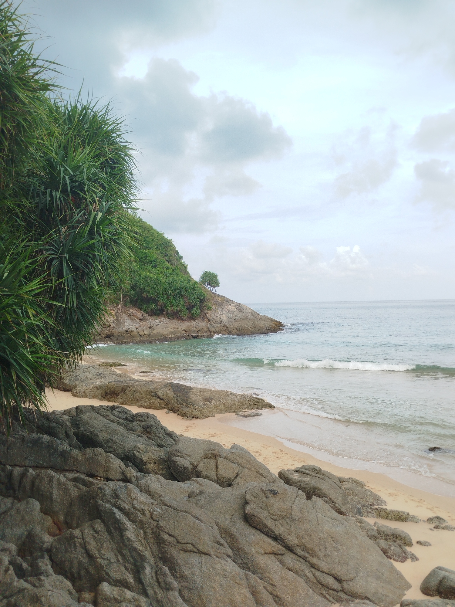 The image shows a rocky beach with calm, turquoise water under a cloudy sky. In the foreground, large, grey rocks are scattered on the sand. To the left, lush green foliage overhangs the scene. In the mid-ground, a sandy beach curves around a rocky outcrop covered in vegetation. A few small trees grow on the top of the outcrop. The background is dominated by the expansive ocean meeting the hazy horizon under a pale sky filled with soft clouds. The scene appears to be a natural, undeveloped coastal area, likely within Rawai, Thailand, given its tropical vegetation and rock formations. There are no people or animals visible in the image.