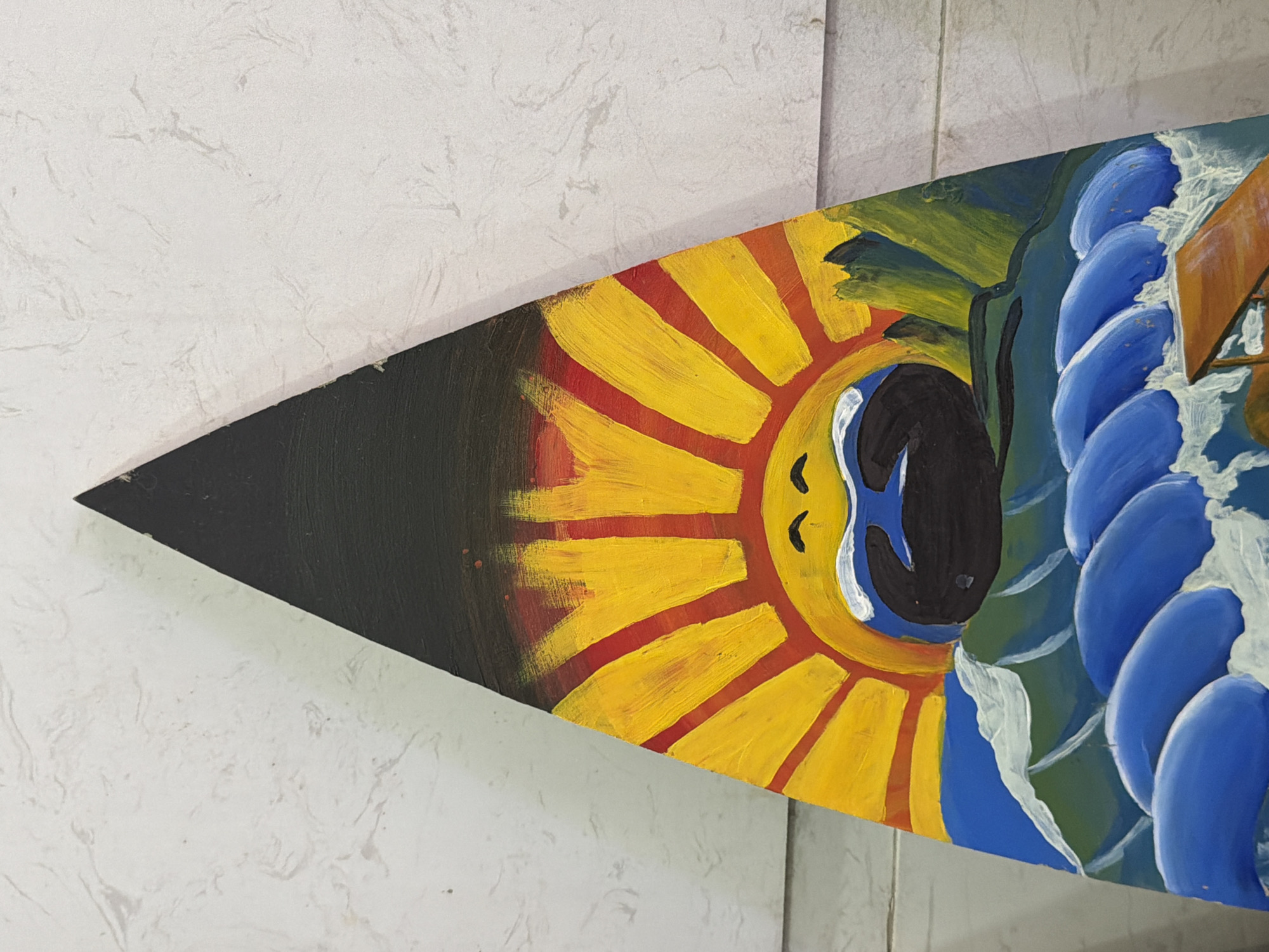 The image shows a triangular piece of wood painted with a depiction of a sun, ocean, and mountains. The sun, with a face, is bright yellow with red rays and is positioned above a dark, curved shape that appears to be a mouth. Below the sun are green mountains. The lower part of the painting features blue waves with white foam. The painted wood is leaning against a textured, light-colored wall. No specific location in Đà Nẵng, Vietnam can be identified from the image.