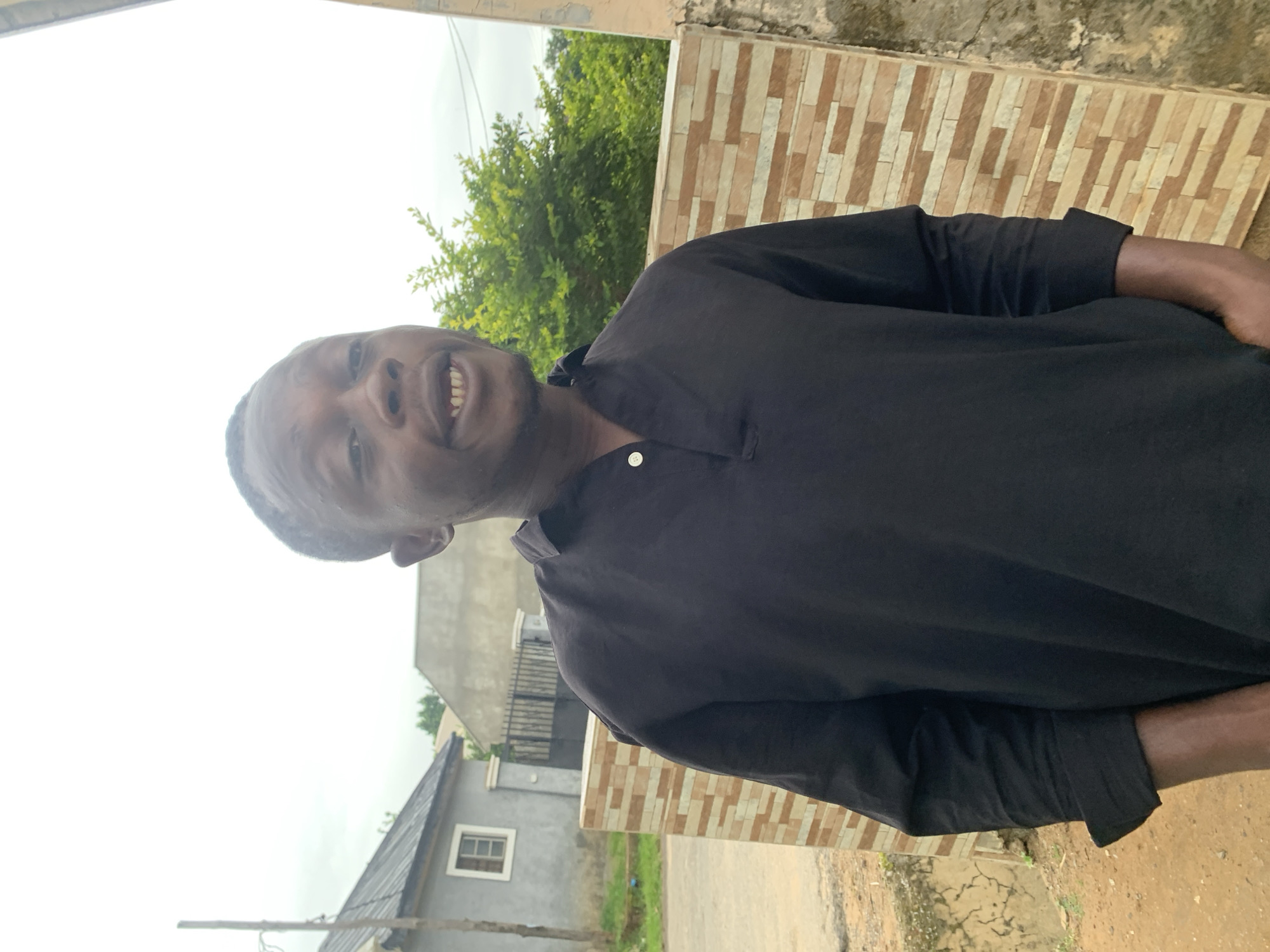 A medium shot shows a Black male standing outside, his body angled slightly to the right. He has a short haircut and his mouth is open, revealing his teeth and gums. He is wearing a black, collared shirt with two buttons at the neckline. The background features a light-colored brick wall to his right, and a grey house with a window on the left. Green trees and foliage are visible behind him. The ground is a dirt path. The sky is overcast. The location is likely a residential area within the city of Abuja, Nigeria.