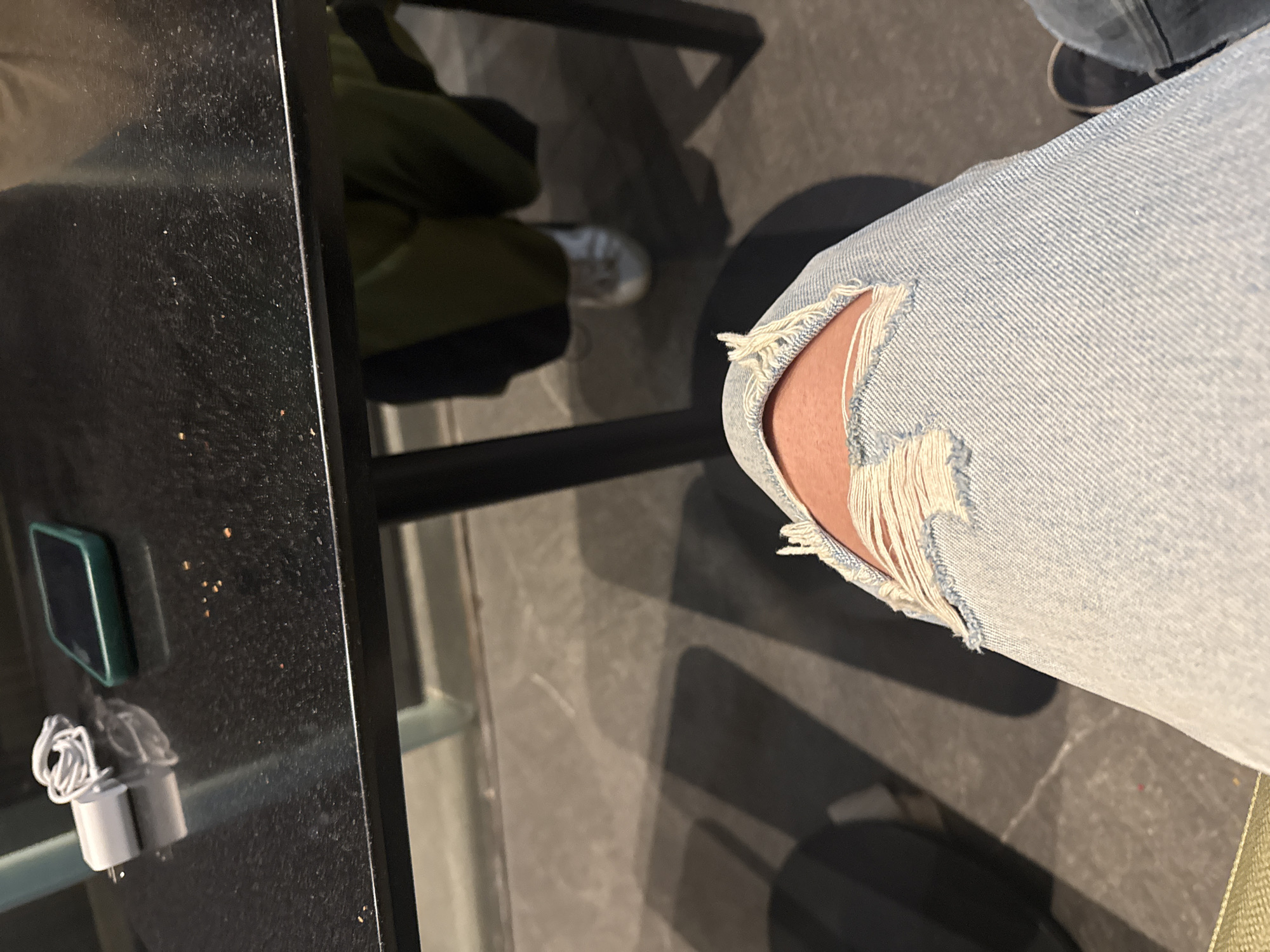 The image presents a partial view of an individual wearing light-wash denim jeans with a distressed, ripped knee. The setting appears to be indoors, near a dark-colored table with electronic accessories (likely a phone charger and a small device) present. The floor is tiled in a neutral gray tone, and a portion of another person, dressed in dark jeans and wearing a dark-soled shoe, can be seen in the background. The rest of the person is wearing green clothing and white shoes. The setting lacks specific identifiable details to place it within Heroica Puebla de Zaragoza or any other specific location.
