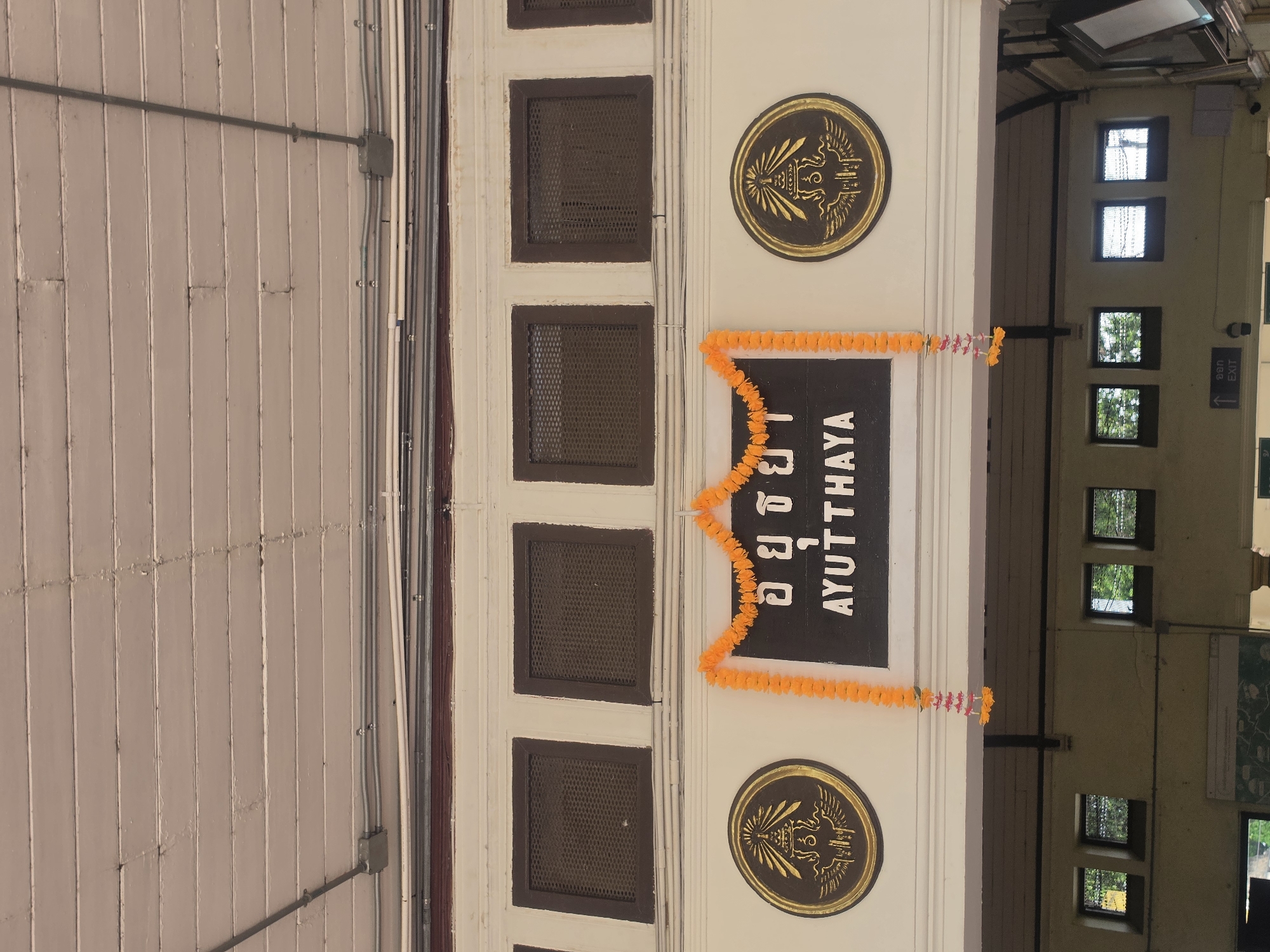 The image shows a building in Phra Nakhon Si Ayutthaya City Municipality, Thailand, with a white facade and dark brown trim. The word "AYUTTHAYA" is written in English and Thai characters on a dark background in the center of the building. There are two gold circular emblems above and below the sign. An orange garland hangs around the sign. To the right of the facade, there's a glimpse into the interior of the building with a view of windows and an "EXIT" sign. The facade has rows of small, square, dark-colored vents. The leftmost part of the image shows a wall with electrical conduits and a junction box.