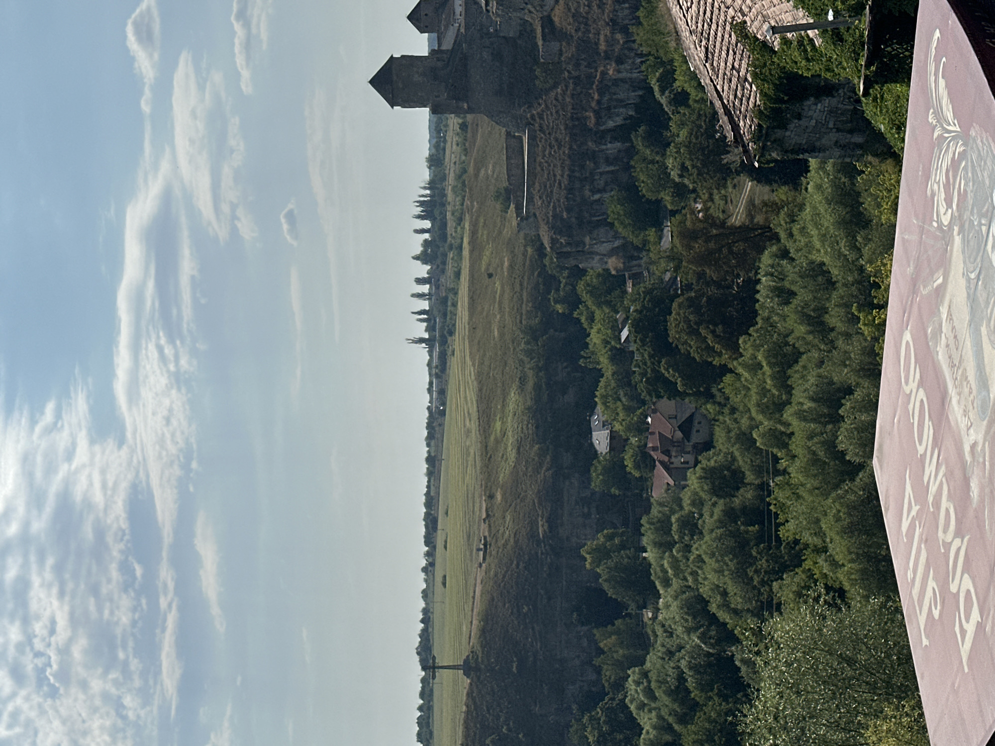 The image shows a landscape view of Kamianets-Podilskyi, Ukraine, from an elevated vantage point. In the foreground, a portion of a table covered with a patterned cloth is visible. The cloth is pinkish-red with white lettering that appears to be in Cyrillic script.
The mid-ground features a lush green valley filled with trees, interspersed with a few buildings with red-tiled roofs. The terrain then rises into a steep, rocky cliff face on which part of the Kamianets-Podilskyi Castle complex is visible. The castle structures, made of stone, include turrets and walls, indicative of historical architecture.
Beyond the valley and castle, the landscape transitions to rolling hills covered in green fields, with a prominent monument resembling a cross on a tall pedestal situated on one of the hills. A line of trees runs along the horizon, and the sky is partly cloudy with a pale blue hue. The overall impression is a scenic view of the historical city and its surrounding natural environment under a daytime sky.