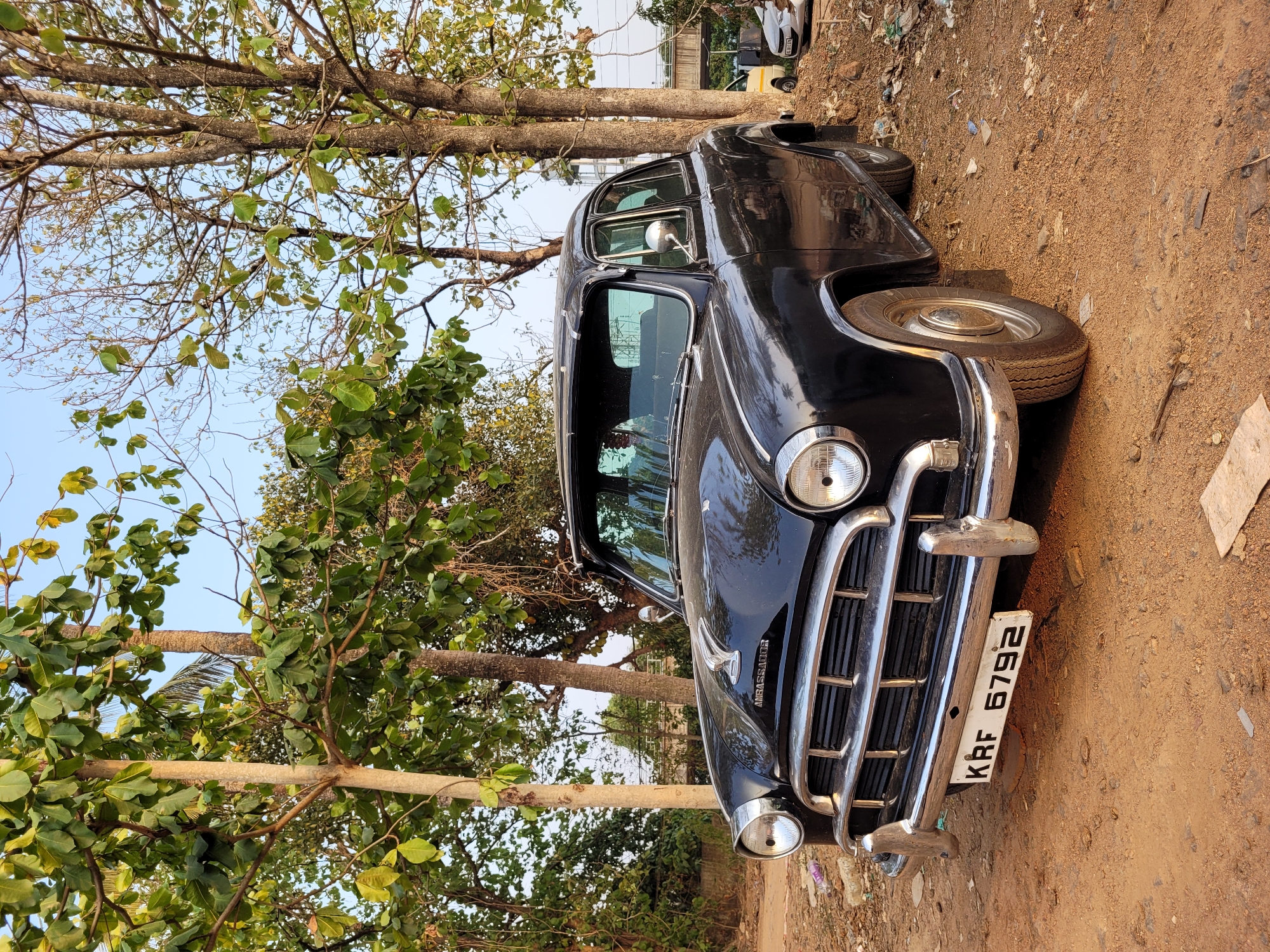 The image shows a black vintage car parked near trees on a dirt surface. The car has a chrome grill and visible headlights. The license plate reads "KRF 6792". The location appears to be a roadside area with vegetation. The environment includes trees, dirt, and sky in the background. The city and state are unknown.