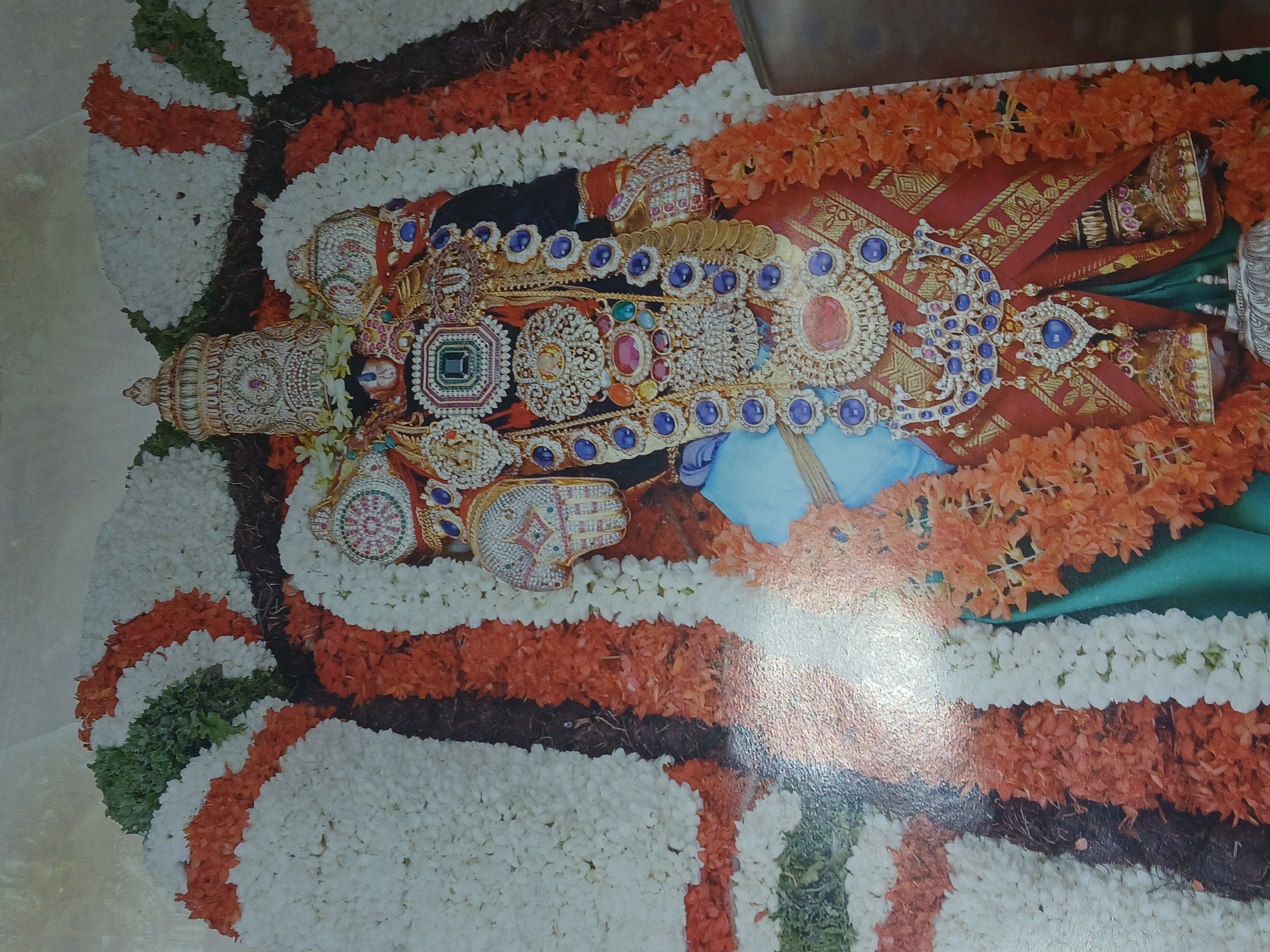 The image depicts a religious scene, likely a deity, adorned with elaborate jewelry, including necklaces and head ornaments, resting on a bed of flowers. The flowers form a patterned border around the deity, featuring white, orange, brown, and green floral arrangements. The deity's attire is predominantly red and gold. Based on the style of the decoration and jewelry, this could be from a Hindu temple in Bengaluru, India. The reflection present in the image suggests that this might be a photograph of the scene.