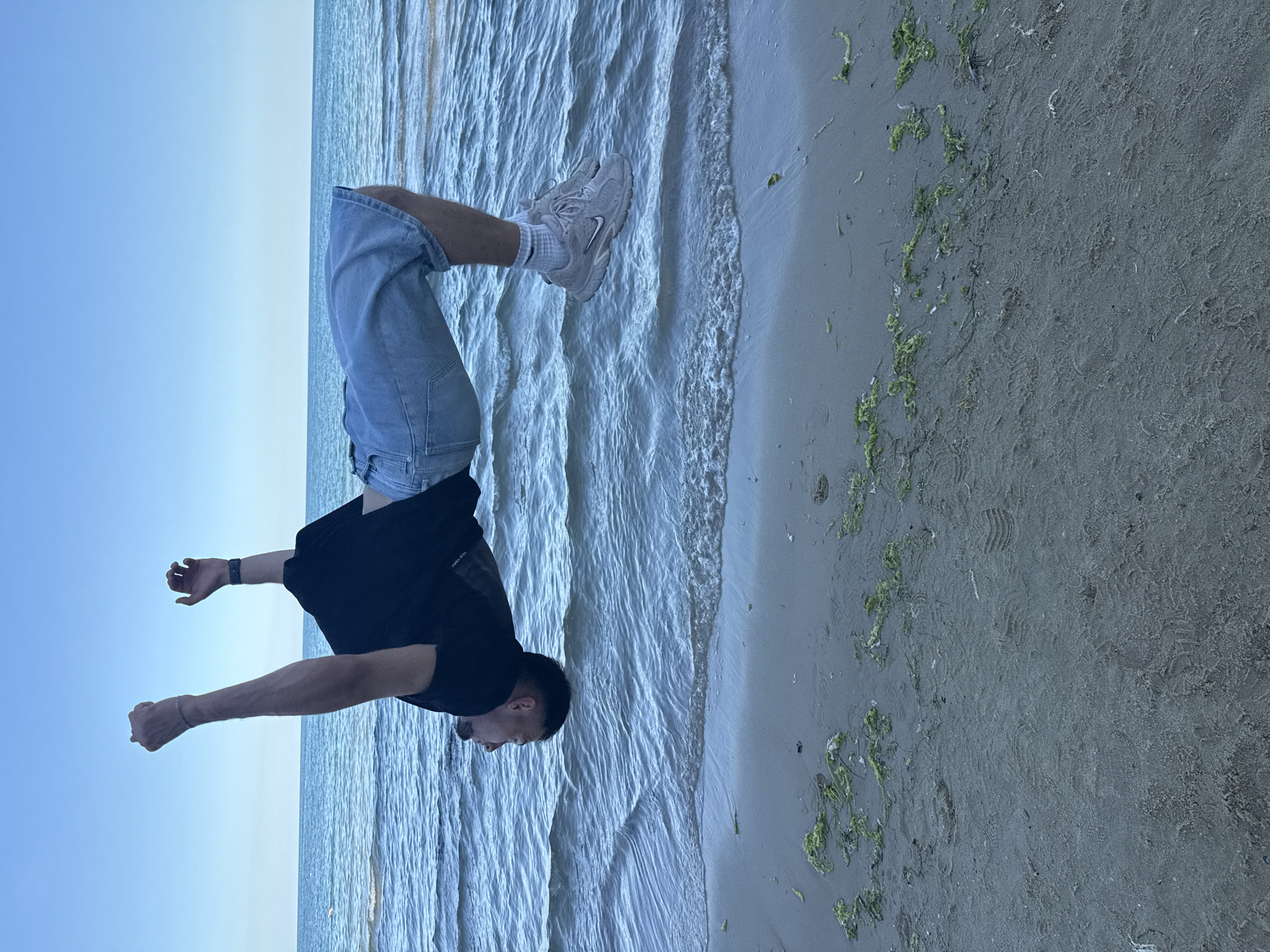 This media file depicts a man performing a handstand on a beach. The man is wearing a black t-shirt, light-wash denim shorts, white socks, and light-colored athletic shoes. His arms are extended, supporting his body upside down. The background features the calm sea with gentle waves lapping against the sandy shore. Patches of green seaweed are scattered on the sand. The sky is a clear, light blue, indicating it is daytime, likely late afternoon or early evening given the soft lighting. The scene conveys a sense of physical activity and enjoyment of the outdoor environment. No visible text is present in the image. The overall impression is one of leisure and athleticism in a seaside setting.