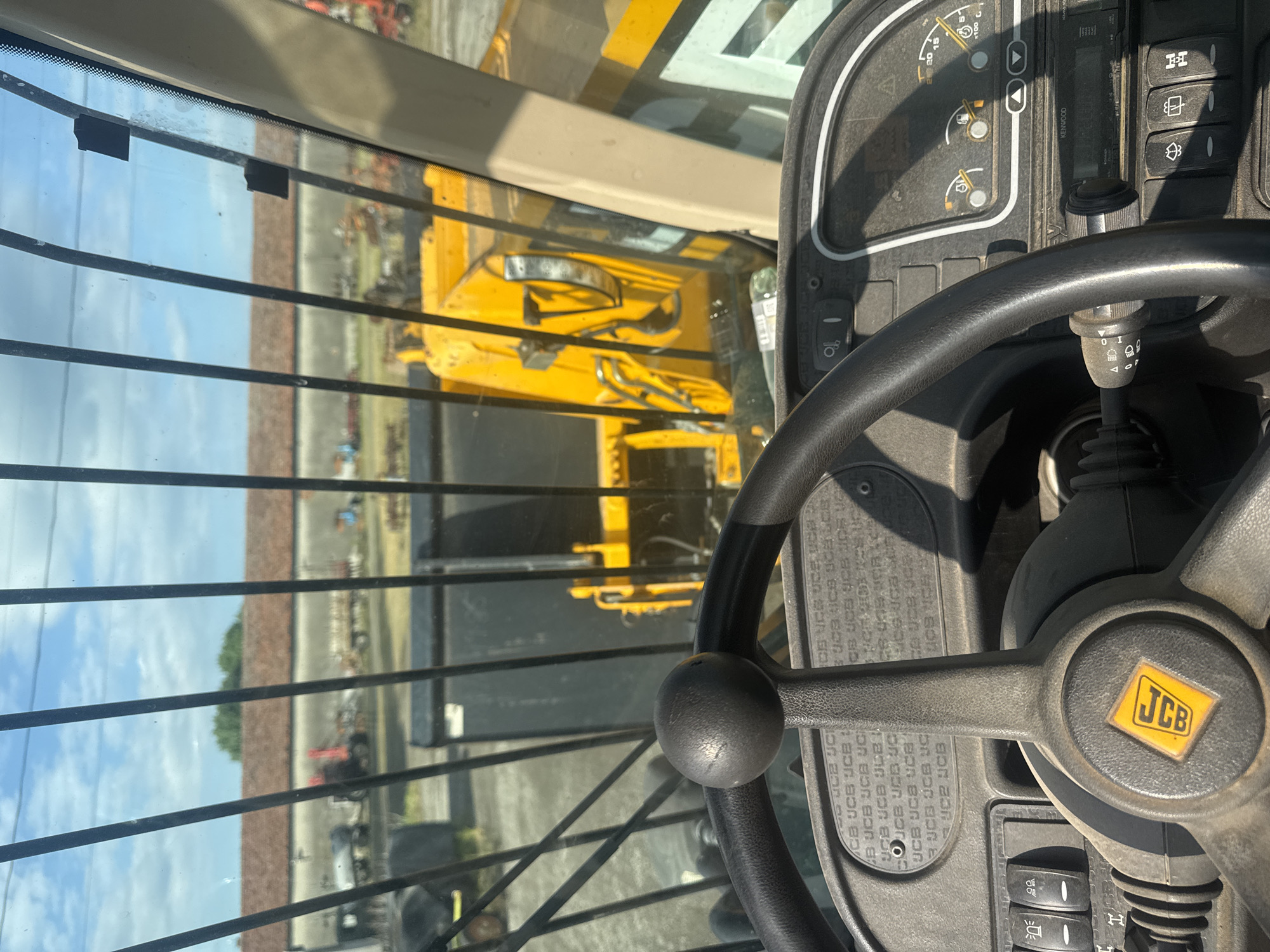 The image presents an interior view from the cabin of a JCB construction vehicle. A steering wheel bearing the JCB logo dominates the foreground, with a dashboard and various controls visible behind it. The view through the cabin window shows another yellow construction vehicle, likely also a JCB, parked outdoors on what appears to be a lot or storage area. A fence or barrier is discernible in the background, along with hints of other vehicles and structures. The OCR analysis confirms the presence of the JCB branding and also identifies a "KENWOOD" component, likely a radio or audio system. The specific location in Sarnevo, Bulgaria, is not discernible from the visual information provided.