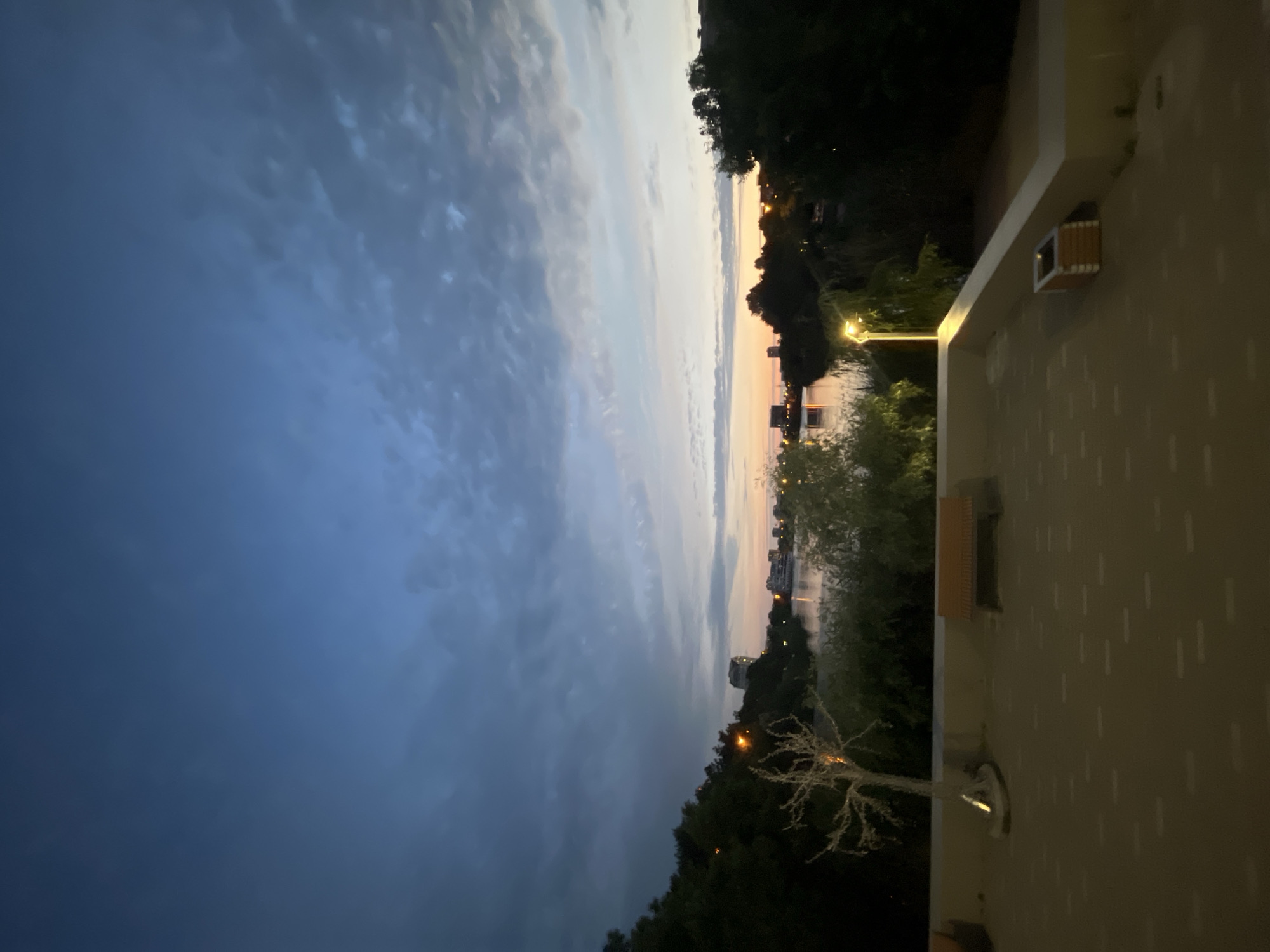 The image depicts a scenic view of a body of water at dusk, likely a river or lake, in the city of Krasnodar, Russia. The sky is a deep blue with scattered clouds, transitioning to lighter hues of orange and yellow near the horizon, indicating the sun has recently set. The water reflects the dim light, with a few buildings and lights visible on the opposite bank.
In the foreground, a paved area with a patterned surface suggests a public space, possibly a promenade or park. Several elements are present on this paved area:
- A bare, tree-like sculpture stands to the left, illuminated by a nearby lamppost.
- A wooden bench is positioned centrally, offering a place to sit and enjoy the view.
- To the far right, a small, rectangular planter box is visible.
The overall atmosphere is serene and tranquil, characteristic of an evening by the water in an urban setting. The lighting suggests it is twilight, with artificial lights from the lampposts beginning to illuminate the foreground. No people are visible in the image.