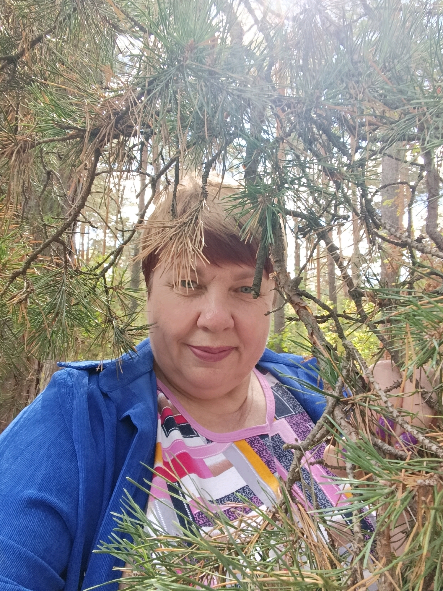 A woman with short, light brown hair and blue eyes is photographed outdoors, partially obscured by pine branches. She is wearing a bright blue corduroy jacket over a multicolored patterned top. The background suggests a wooded environment, likely a park or forest, with tall trees and dappled sunlight filtering through the foliage. The image is likely a selfie, given the angle and framing. There is no clear indication of the specific location within Svietlahorsk, Belarus, beyond it being an outdoor, wooded setting.