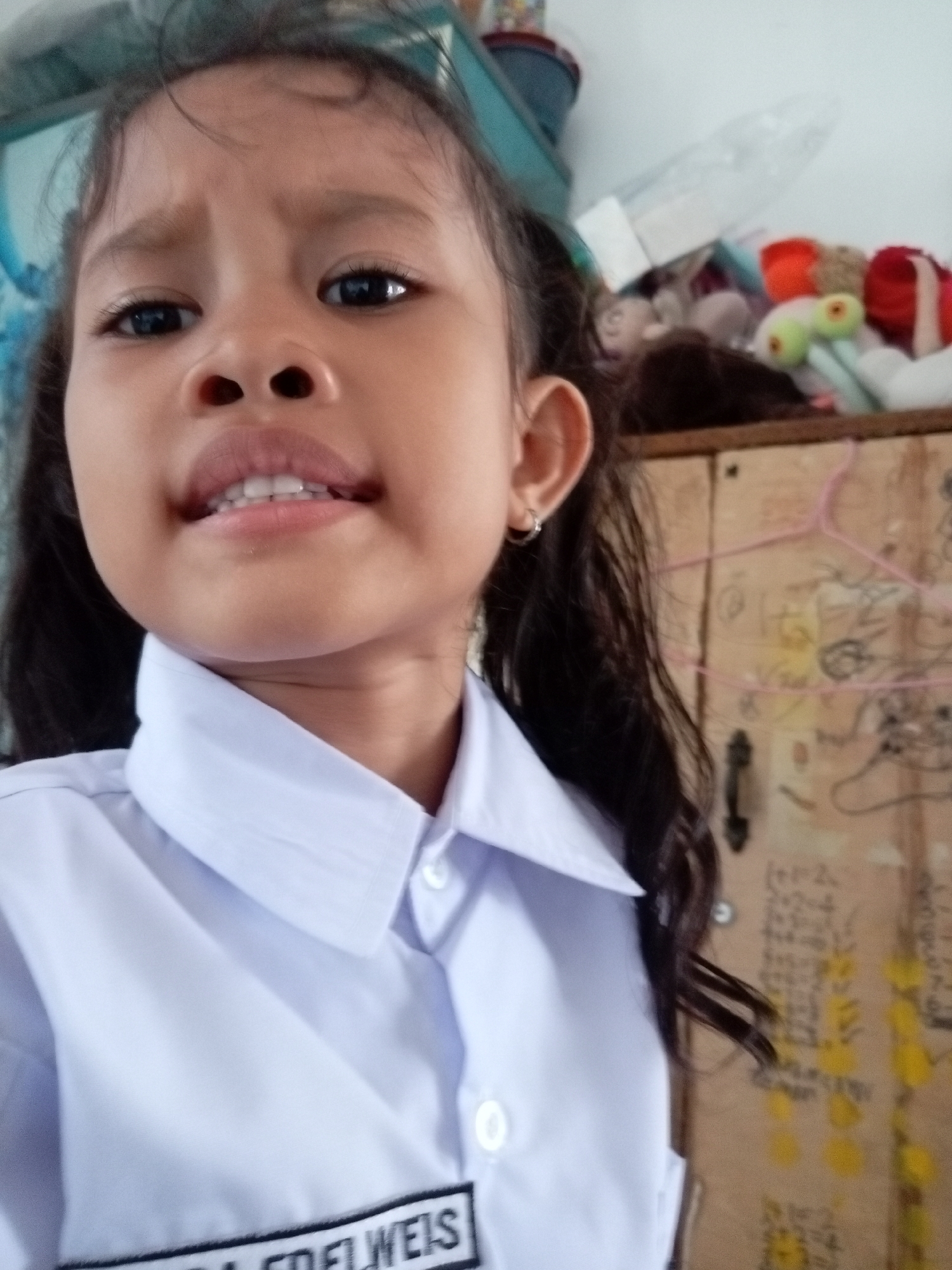 The image is a close-up, selfie-style shot of a young Indonesian girl in Batam, Indonesia. She is wearing a white collared shirt with a name tag that appears to read "EDELWEIS". Her expression is a bit uneasy, with furrowed brows and slightly parted lips revealing her teeth. Her dark hair is pulled back into pigtails, and she is wearing a small hoop earring in her left ear.
In the background, to the right of the girl, is a wooden cabinet with various items on top, including stuffed toys in bright colors. The cabinet door is decorated with what looks like handwritten notes or drawings. The overall setting appears to be indoors, possibly a home or classroom, given the presence of toys and the personal nature of the photograph. The lighting suggests it could be daytime, but no specific time of day can be determined. The weather is not discernible from the image. The focus is primarily on the girl's face and upper body, with the background slightly out of focus.