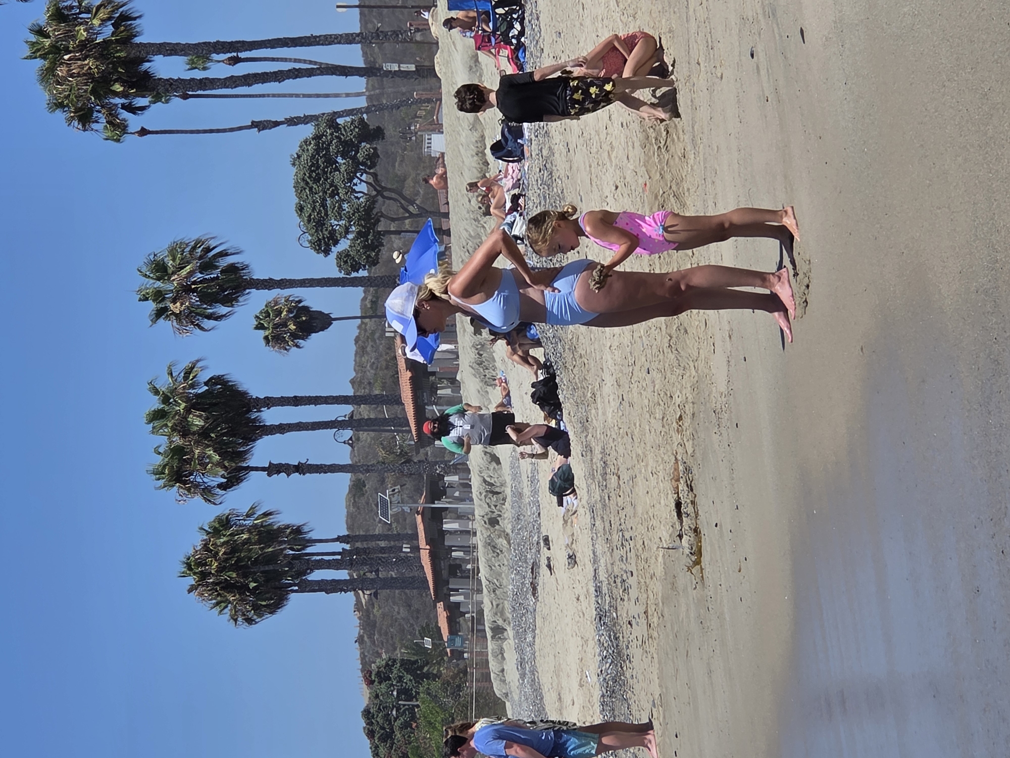 The image depicts a sunny day at San Clemente State Beach in San Clemente, California, USA. In the foreground, a woman in a light blue bikini and a white cap stands with her hands on her hips, looking to the left. Beside her, a young girl in a pink swimsuit holds a piece of sand. To their right, a young boy in a black shirt and patterned swim trunks stands facing away. In the background, several people are scattered across the sandy beach, some lying on towels or sitting in chairs, under palm trees and a blue umbrella. The shoreline features pebbles and dry seaweed. Further back, a row of buildings with orange roofs and solar panels line the edge of the beach, with a hilly, vegetated landscape beyond.