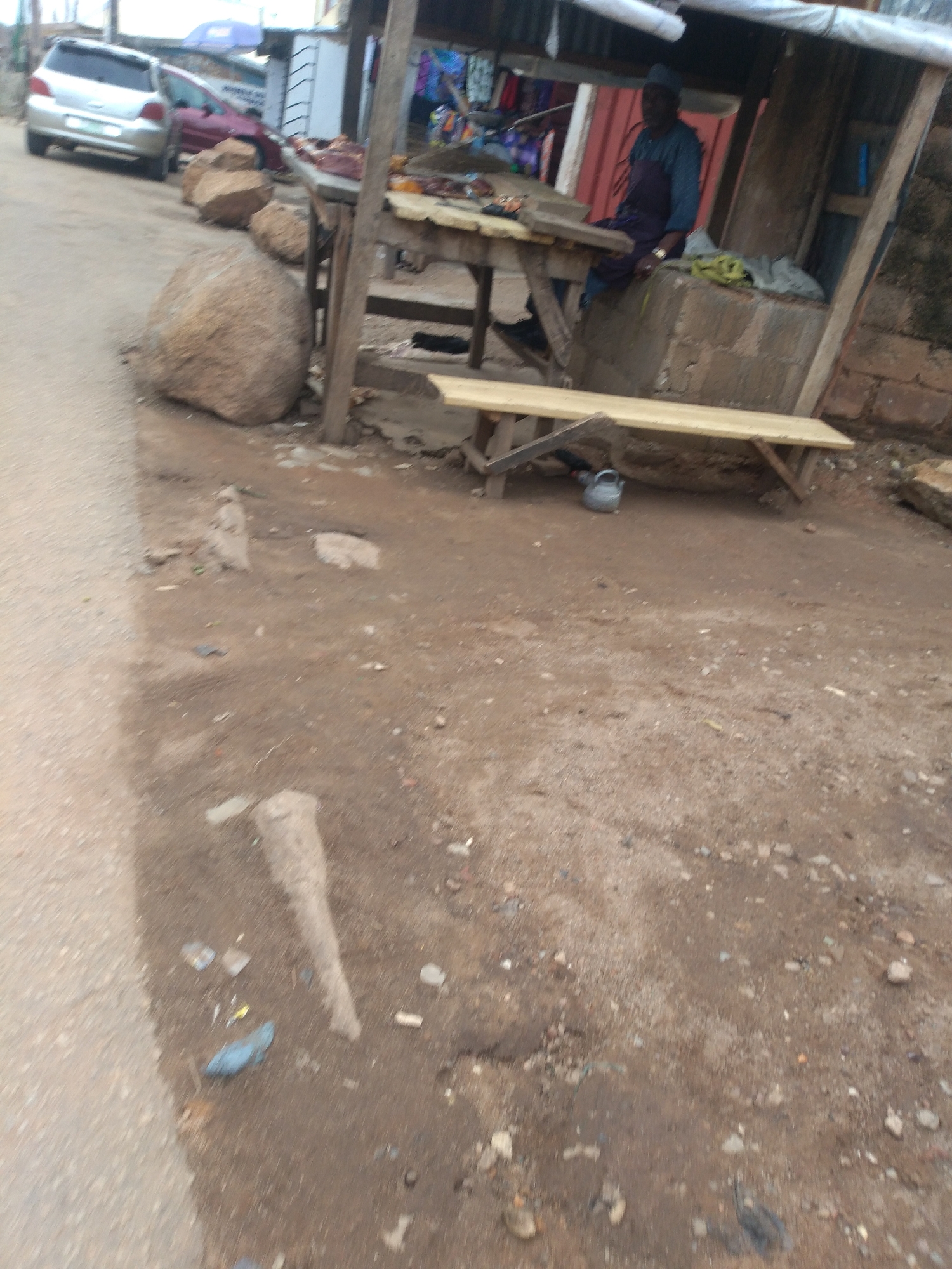 The image depicts a roadside market stall in Jos, Nigeria. A man wearing a blue and purple garment and a cap sits in the stall. In front of the stall is a wooden table displaying what appears to be raw meat. A wooden bench is positioned in front of the table. The ground is unpaved and dusty, with scattered debris. To the left of the stall, a silver car and a red car are parked. Large rocks are placed near the stall and the parked cars. The scene suggests an outdoor commercial area within the city.