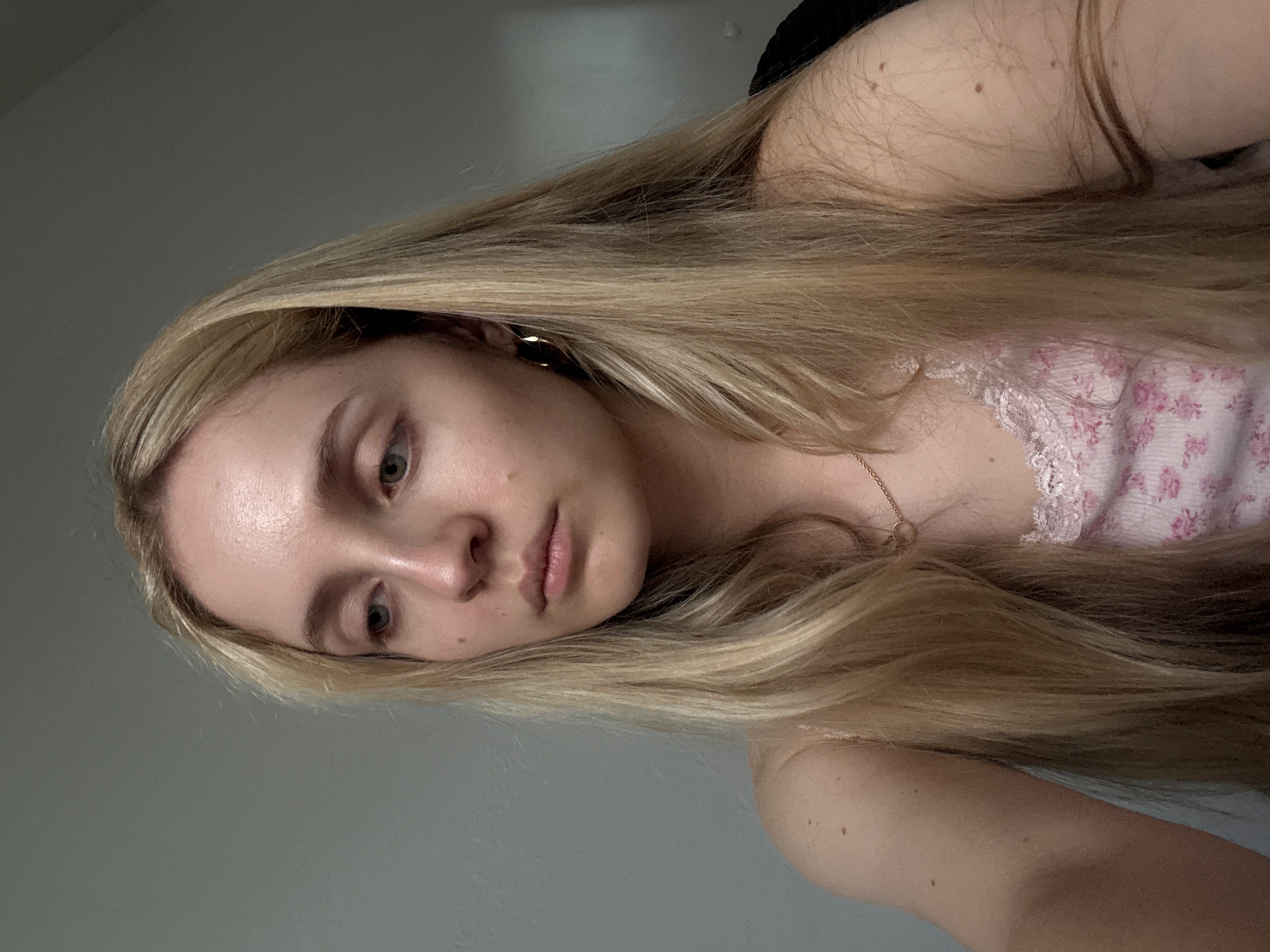 A medium shot shows a young woman with long, blonde hair looking down and to her left. She is wearing a pink camisole with a floral pattern and lace trim. Her left ear is visible and she is wearing a small gold hoop earring. A delicate gold chain necklace is also visible around her neck. The background is a plain, light gray wall. The lighting appears to be soft and natural, casting subtle shadows on her face and neck.
