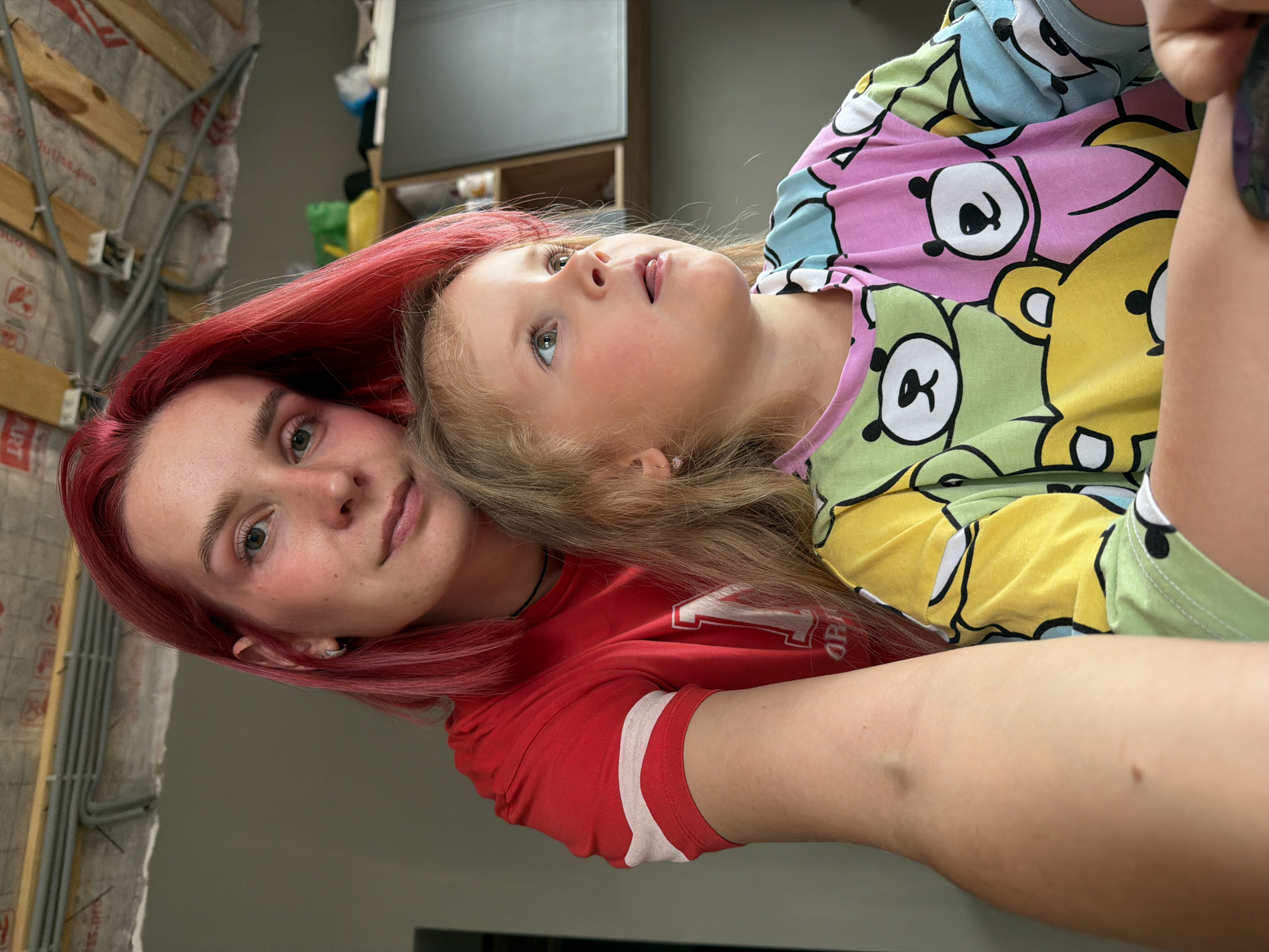 In this medium shot, two individuals are pictured indoors, seemingly in a residential or unfinished construction environment. On the left, a young woman with vibrant red hair, wearing a red t-shirt with white trim, looks towards the camera. Her gaze is direct, and her expression is neutral. Positioned in front of her, slightly to the right, is a young child with blonde hair, also looking to the right, away from the camera. The child is wearing a long-sleeved shirt adorned with a colorful pattern of cartoon bears in shades of green, pink, yellow, and blue. The background features exposed ceiling beams and wiring, indicative of ongoing construction or renovation. Shelving units with various items are visible further back, along with a dark, flat surface that could be a screen or appliance. The overall lighting suggests an indoor setting, likely during the daytime. The image does not provide specific geographic markers to pinpoint the exact location within Chaikovsky, Russia.