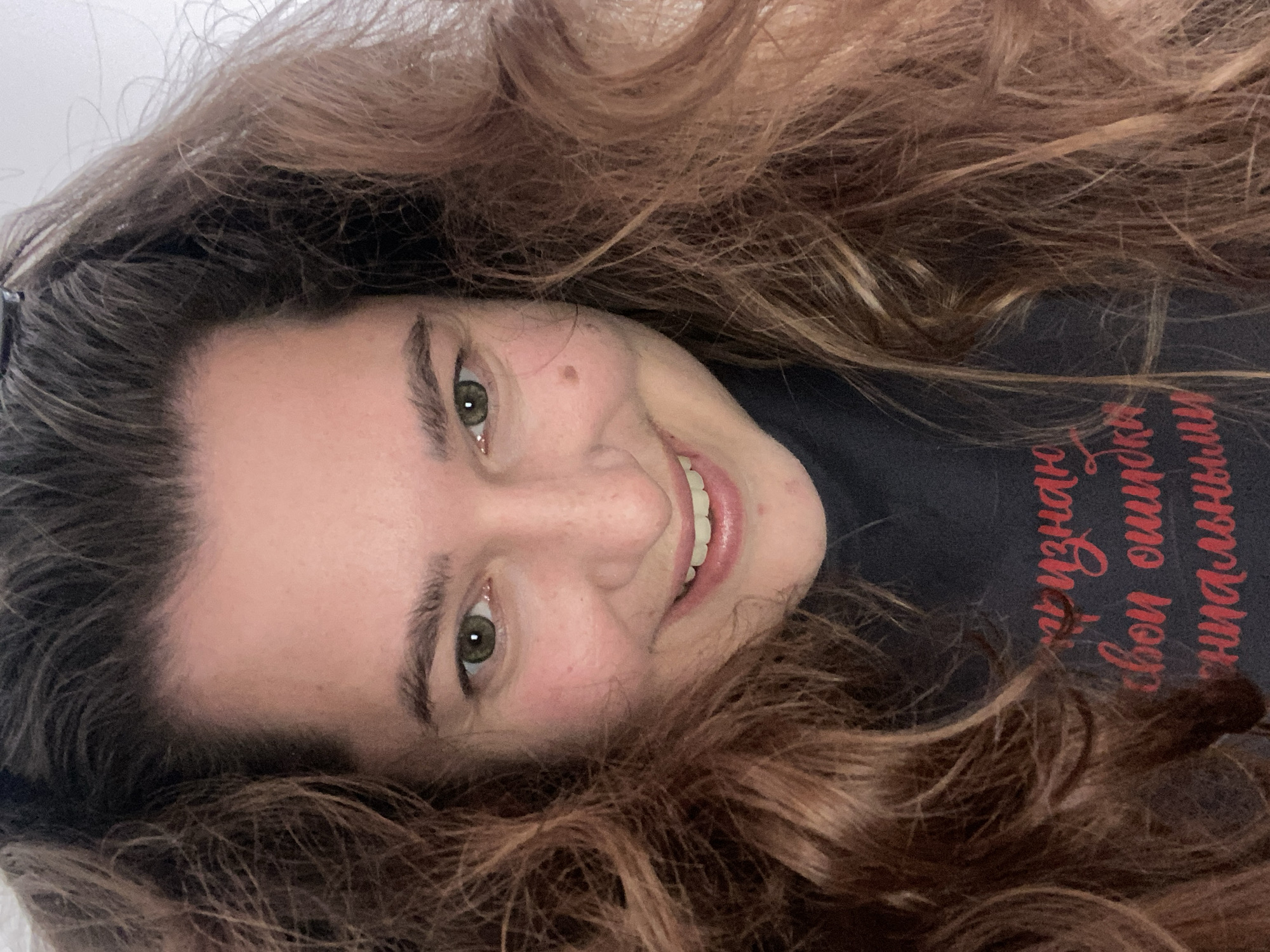 The image is a close-up selfie of a woman with long, wavy brown hair framing her face. She has light skin, green eyes, and a mole on her left cheek. She is smiling with her teeth visible and appears to be wearing a dark-colored shirt with red Cyrillic writing. The background is plain and light-colored, suggesting an indoor setting. The text on the shirt, visible in the lower portion of the image, reads "признаю свои ошибки" which translates to "I admit my mistakes". There are no discernible elements in the image that would allow for the specific identification of a location in the city of Perm, Russia.
